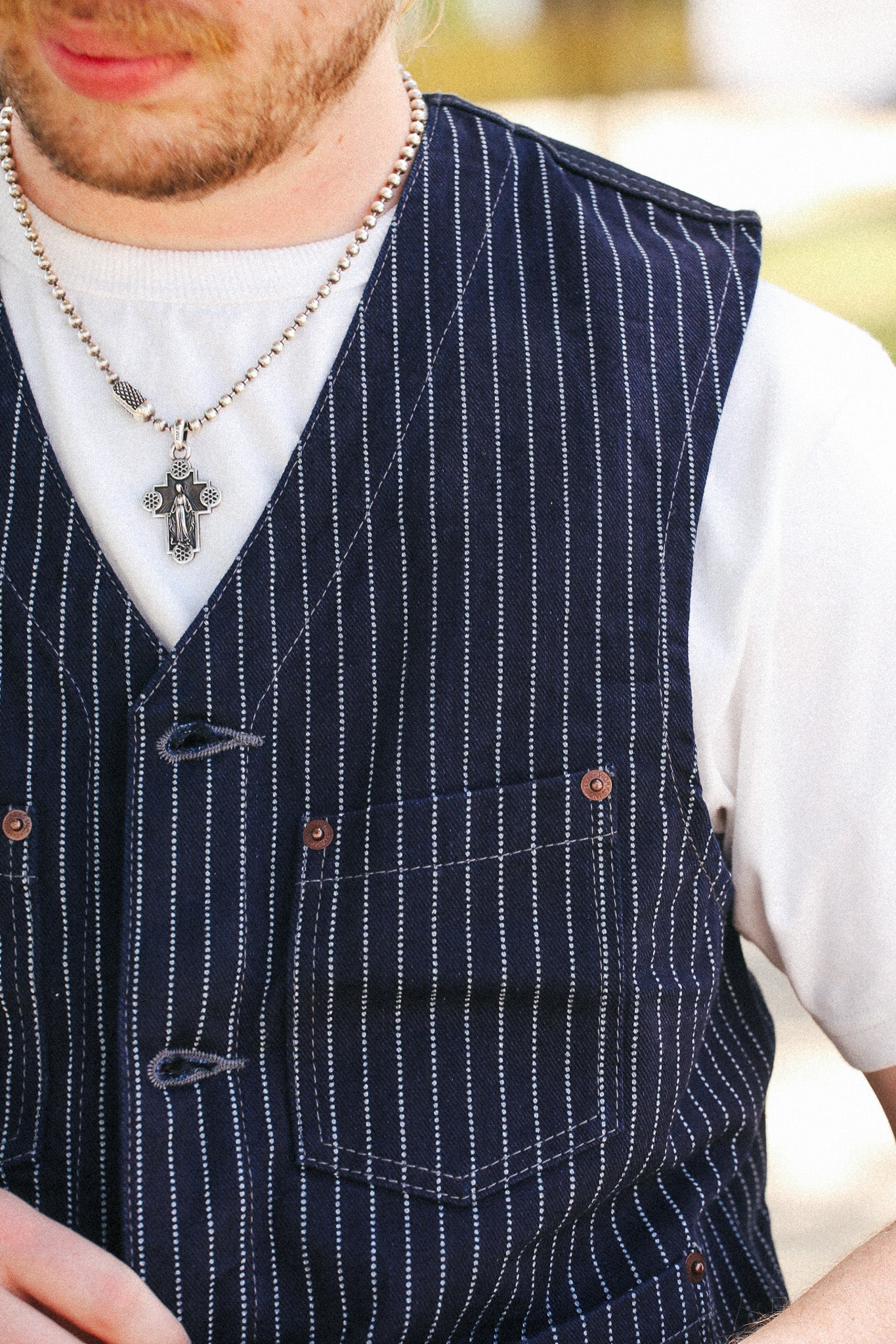 A close-up of a person wearing a navy blue pinstriped vest over a white t-shirt, accessorized with a silver chain necklace featuring a cross pendant.