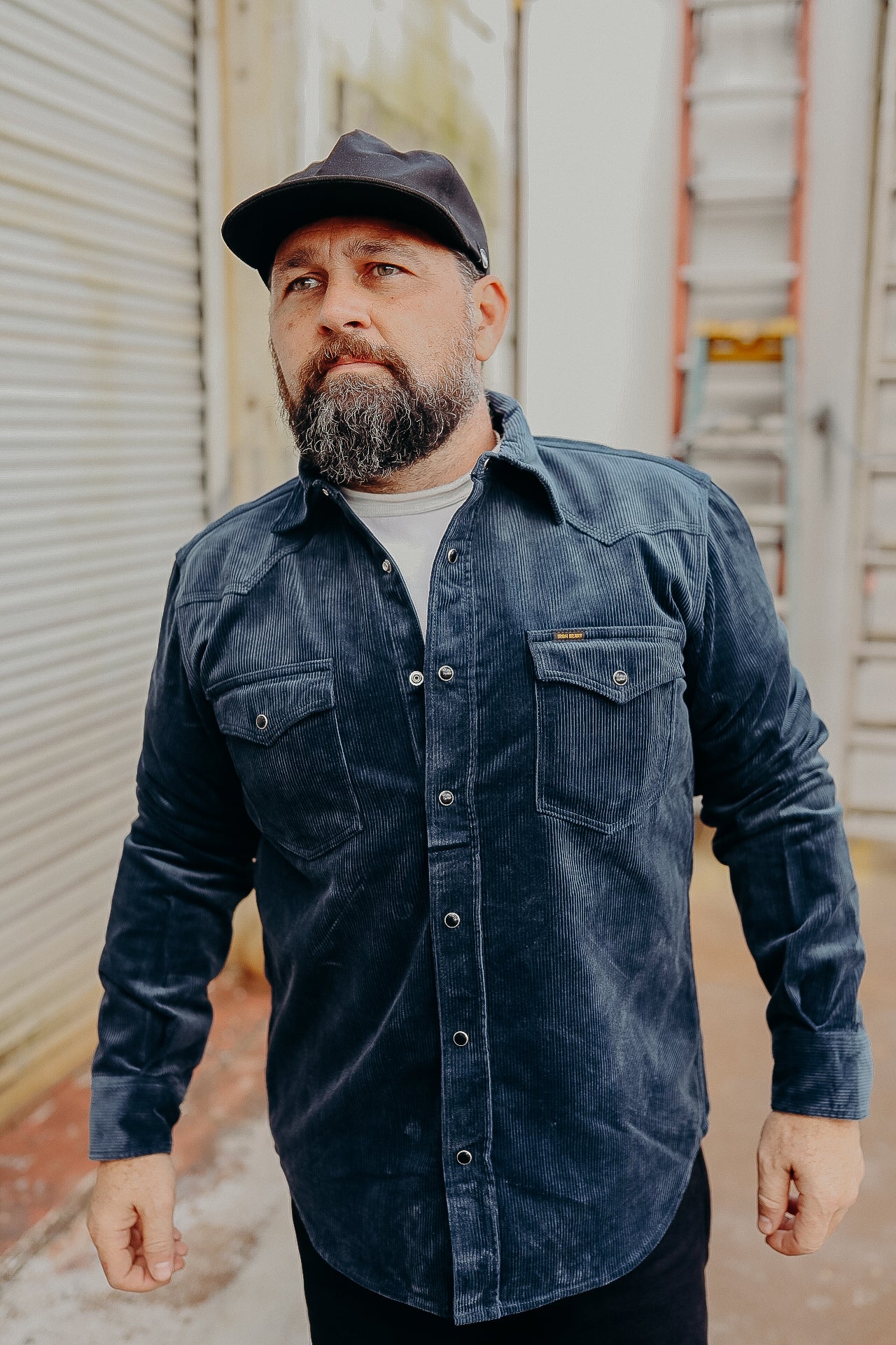 A person wearing a navy corduroy western shirt with pearl snap fastenings, standing in front of a metal gate.