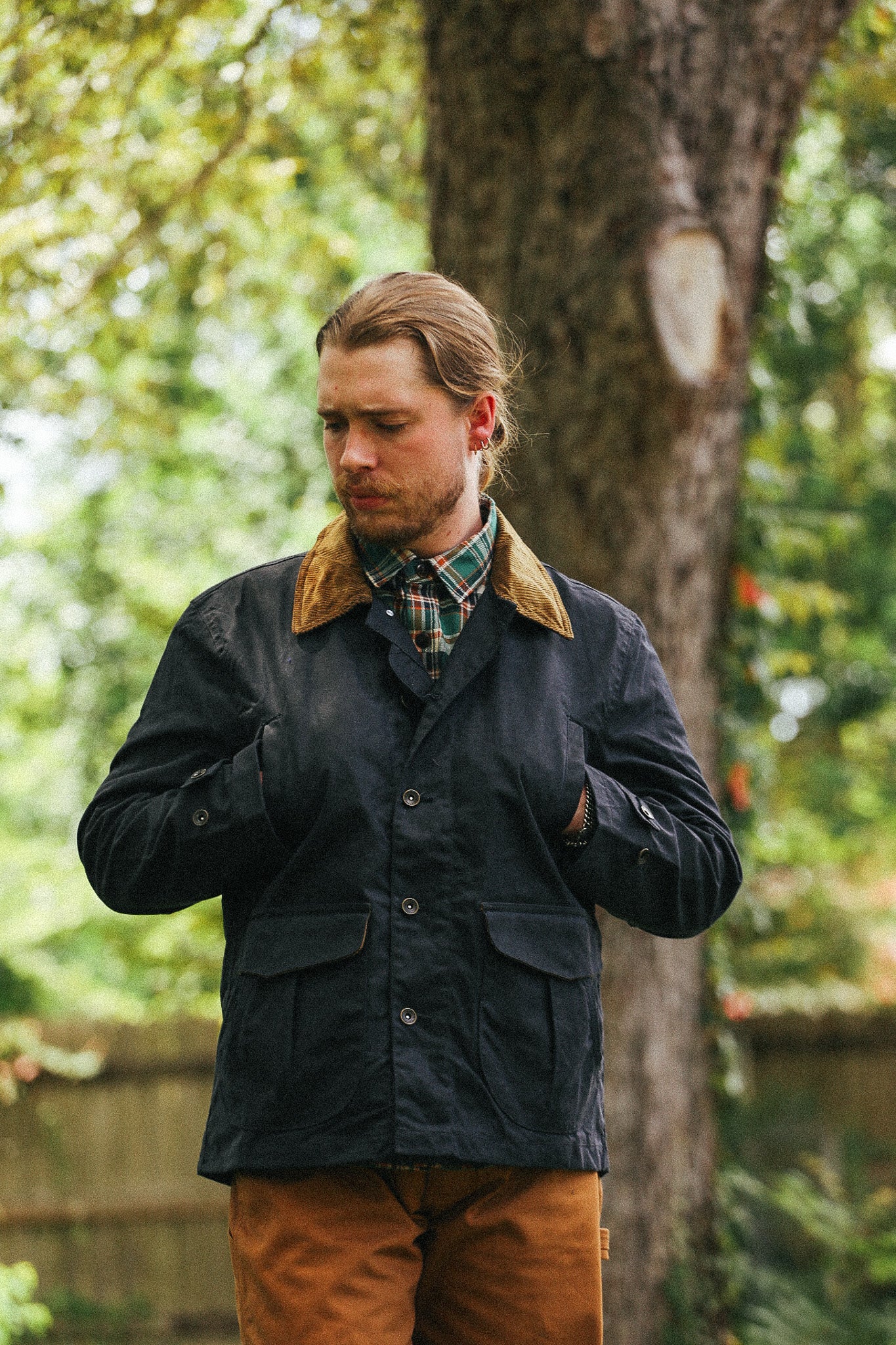 man standing with black jacket and green shirt with hands in pockets against a green natural background 