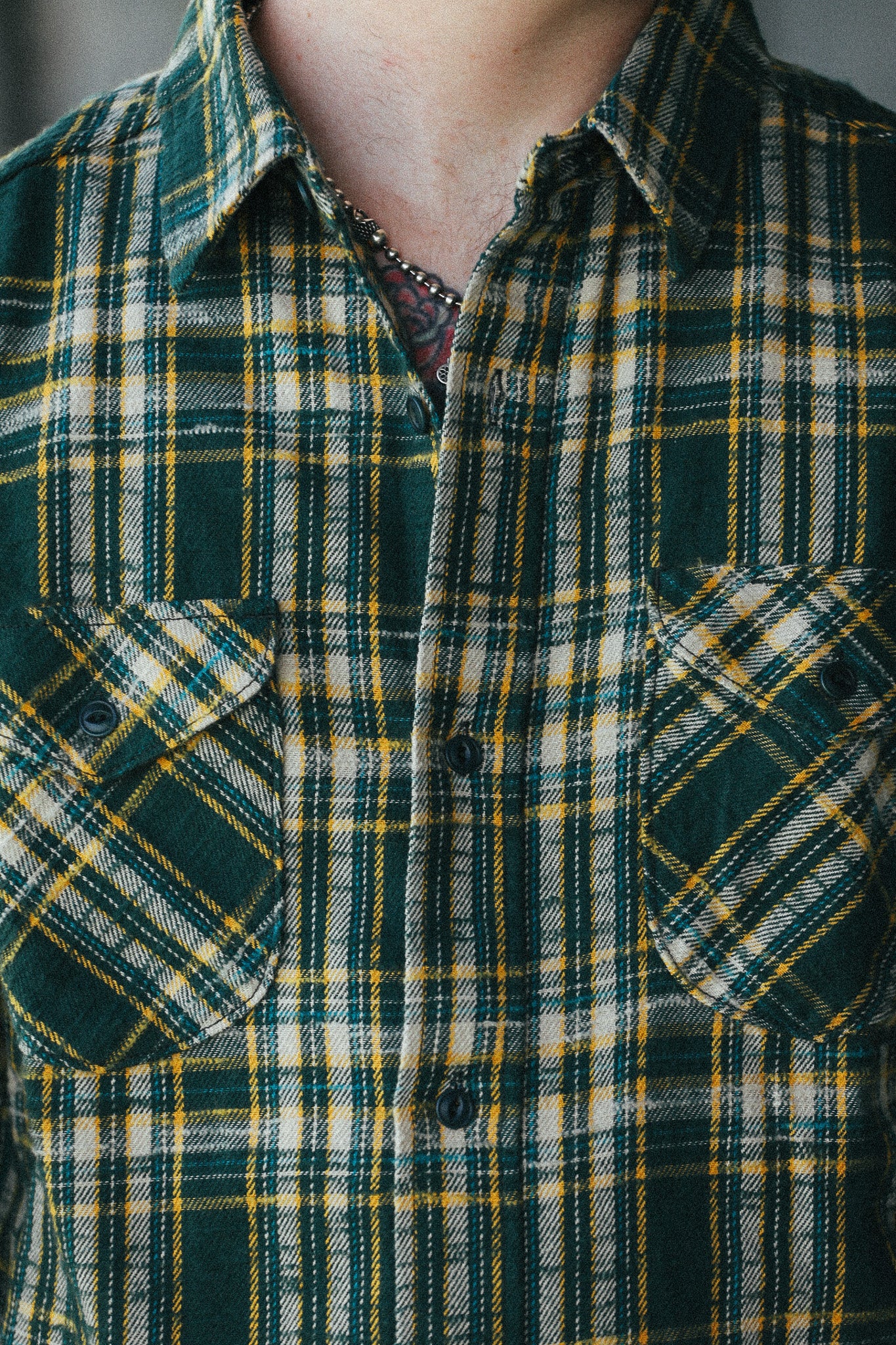 A close-up of a person wearing a green, yellow, and white plaid button-up shirt with two chest pockets.