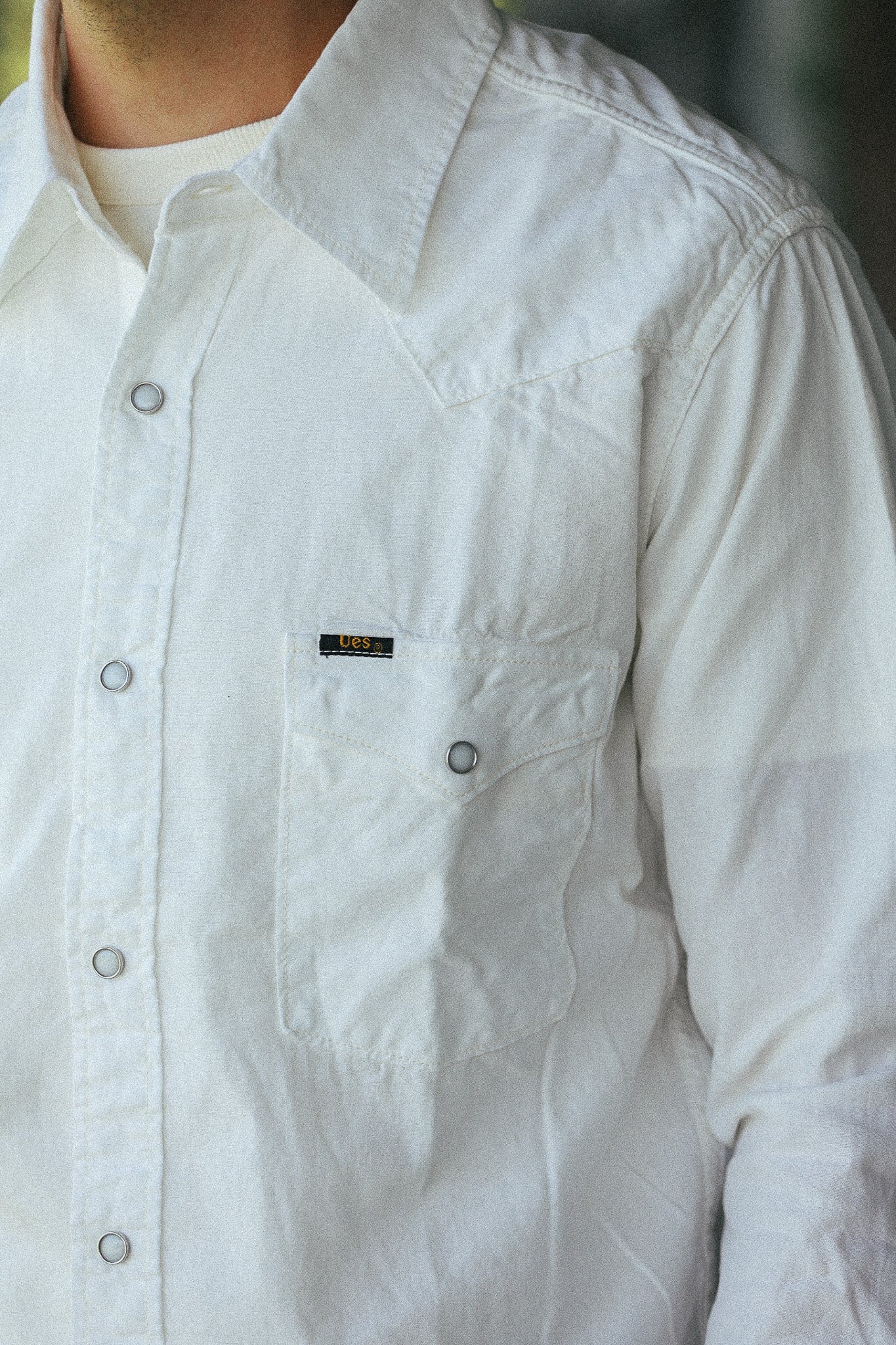 A close-up of a person wearing a white long-sleeve button-up shirt with a chest pocket and a small black brand label.