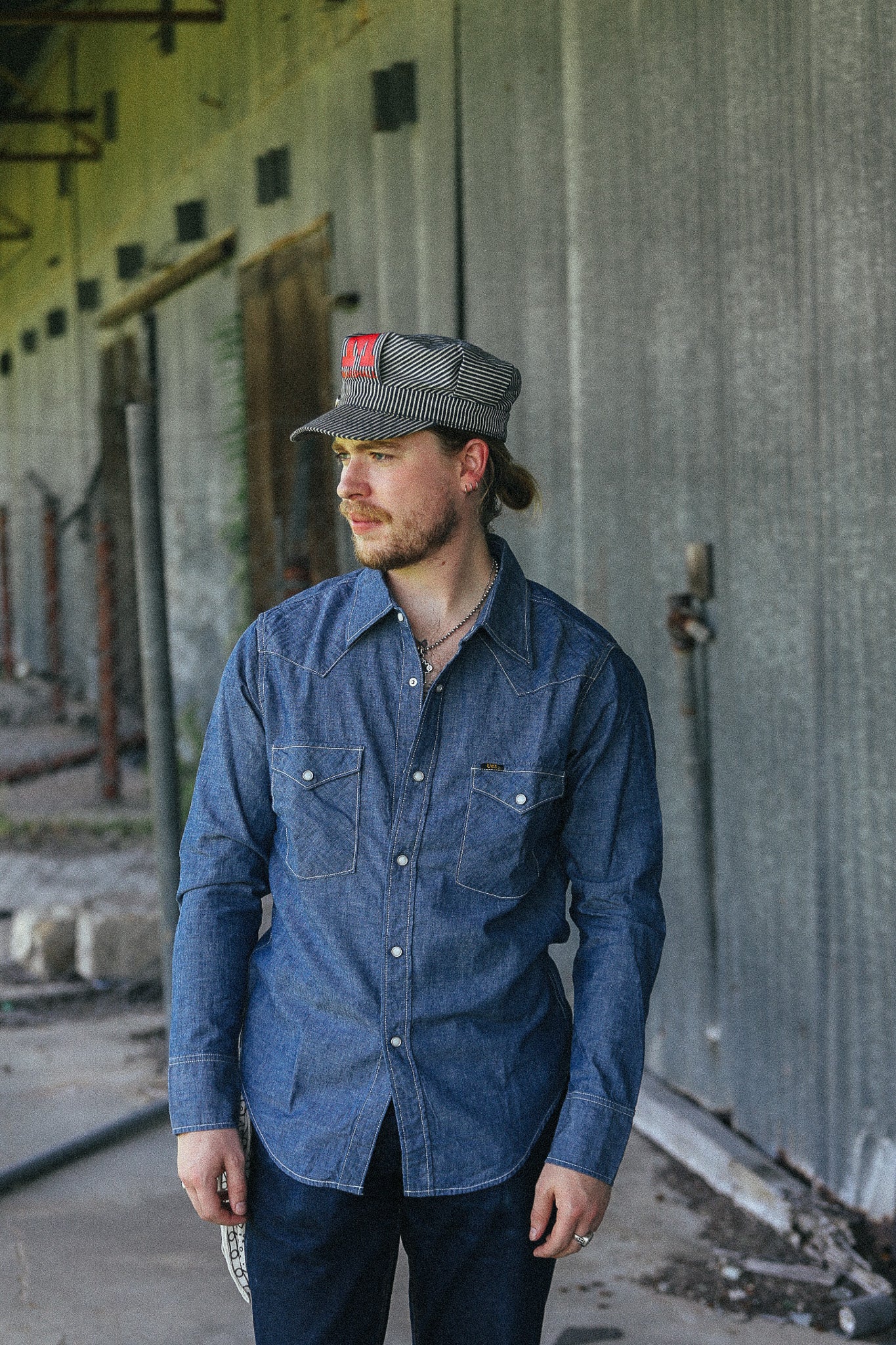 A man wearing a blue denim long-sleeve button-up shirt and a gray patterned cap with a red logo, standing in front of a corrugated metal wall.