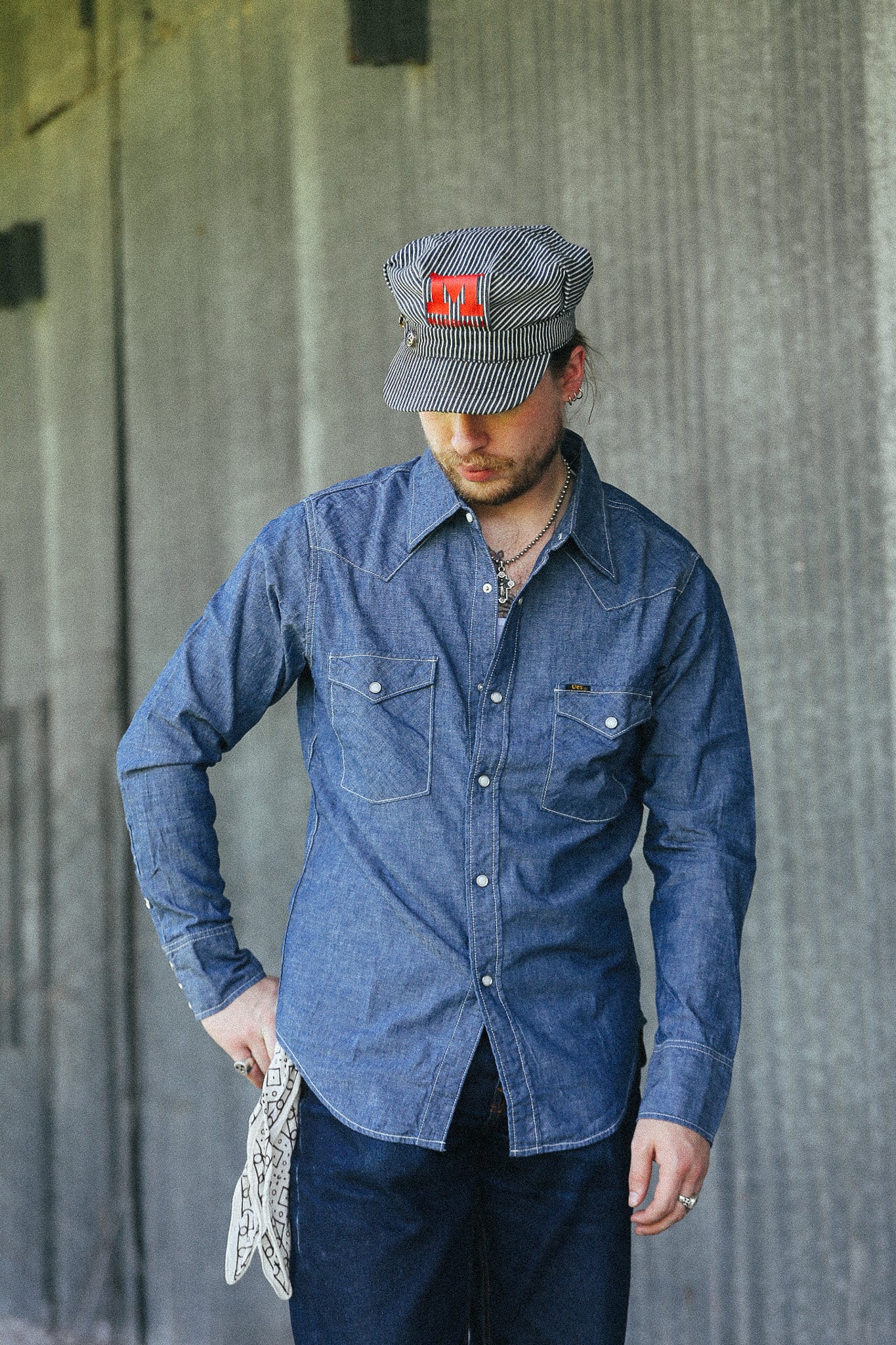 A man wearing a blue denim long-sleeve button-up shirt, dark jeans, a gray striped cap with a red logo patch, and holding a white knit sweater, standing against a textured gray concrete wall.