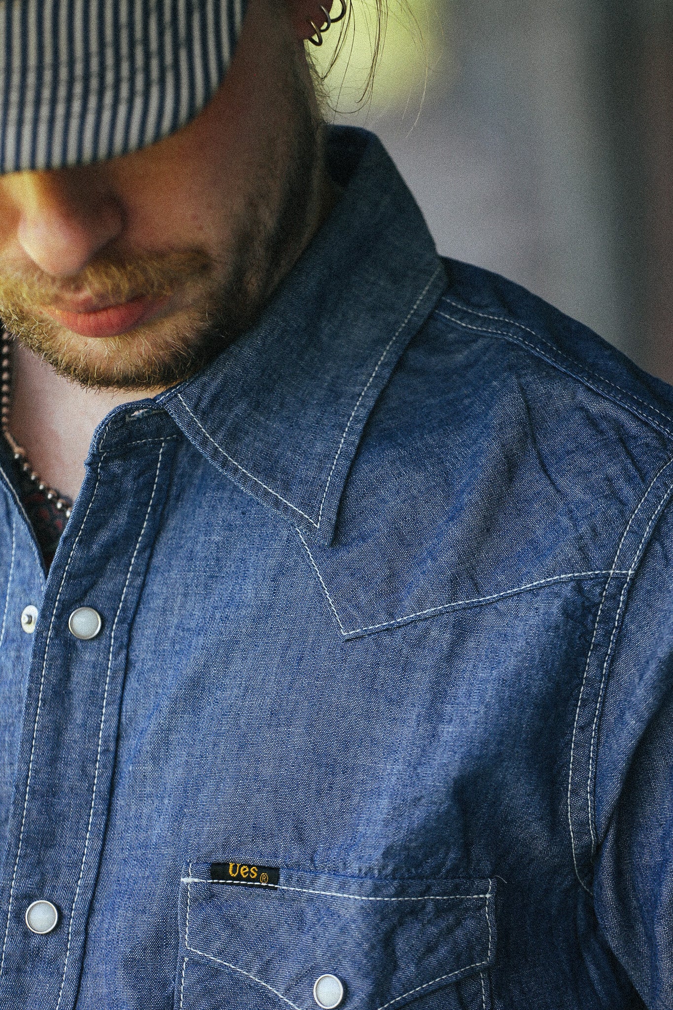 Close-up of a man wearing a blue denim button-up shirt with white contrast stitching and a chest pocket featuring a small yellow and black LEE'S label.
