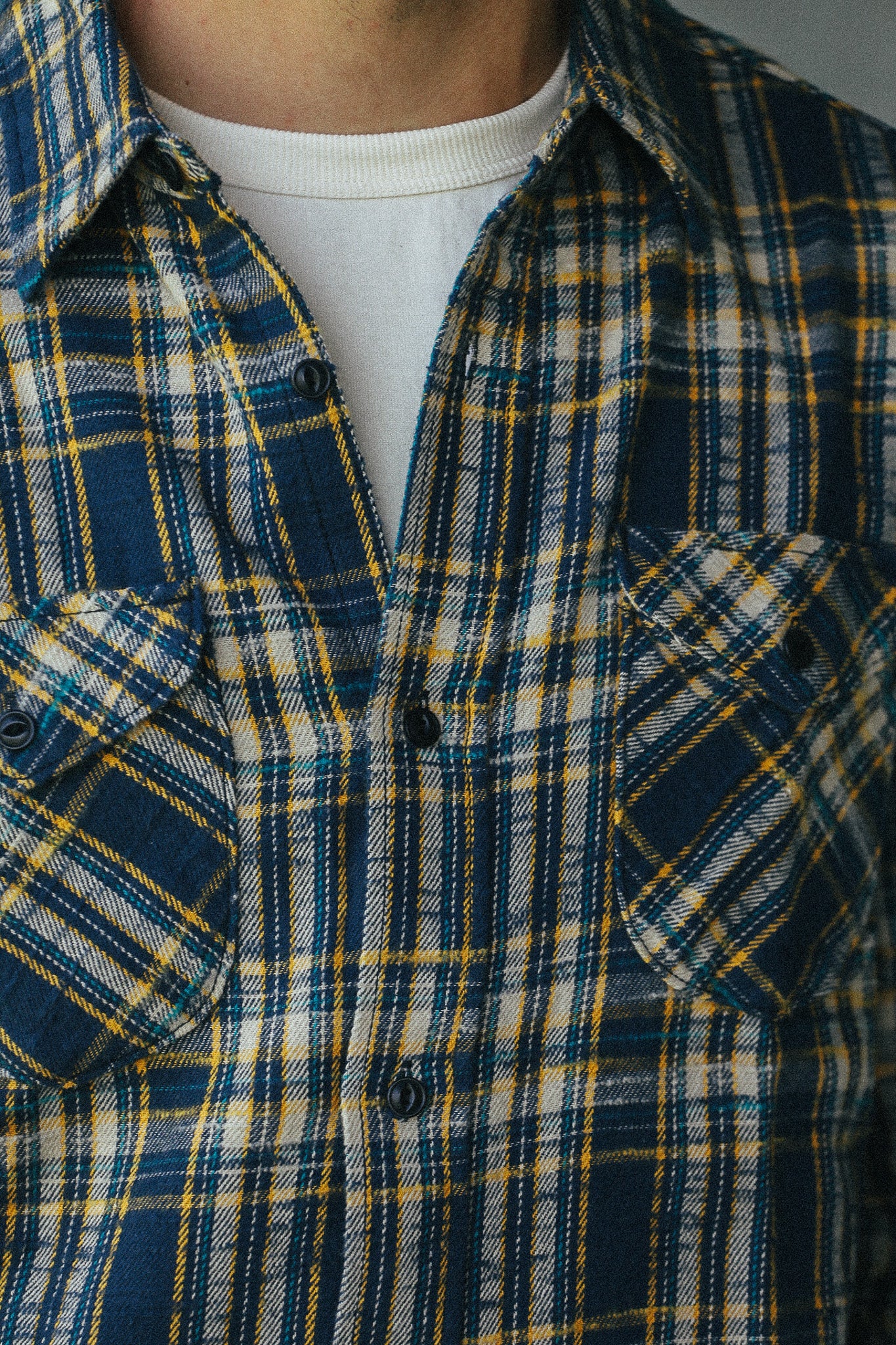 Close-up of a person wearing a blue, yellow, and white plaid button-up shirt over a white crewneck undershirt.