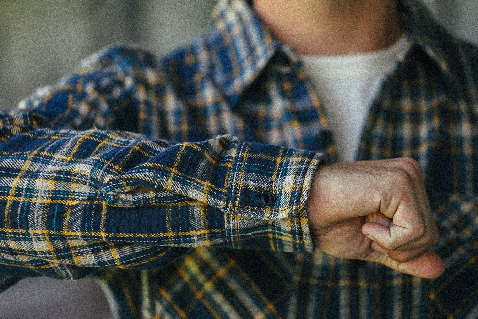 A close-up of a person wearing a blue, yellow, and white plaid flannel shirt over a white undershirt, with their right arm bent forward in a fist.