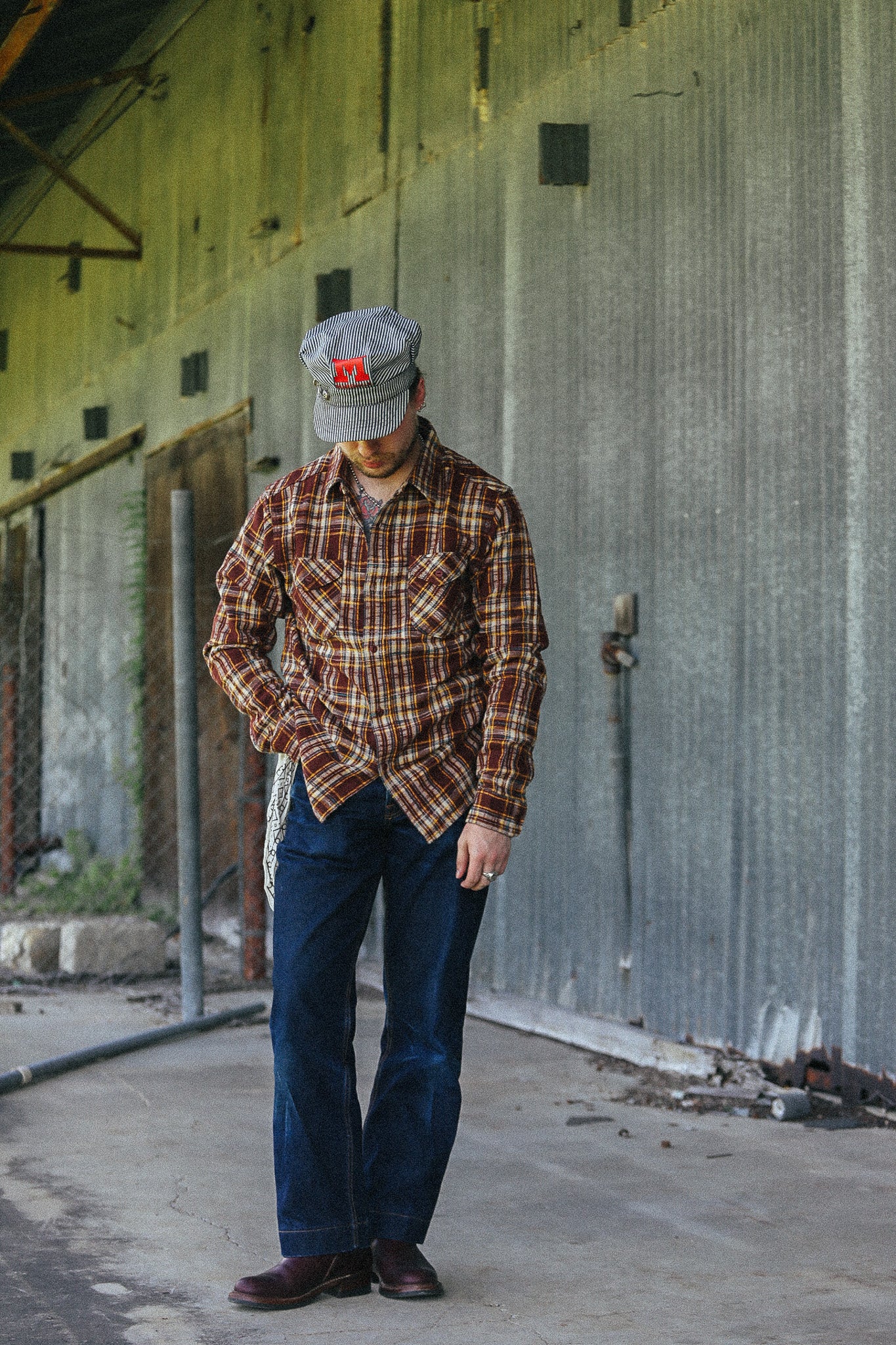 A man wearing a brown and red plaid flannel shirt, dark blue jeans, and a gray cap with a red logo, standing in front of a corrugated metal warehouse wall.