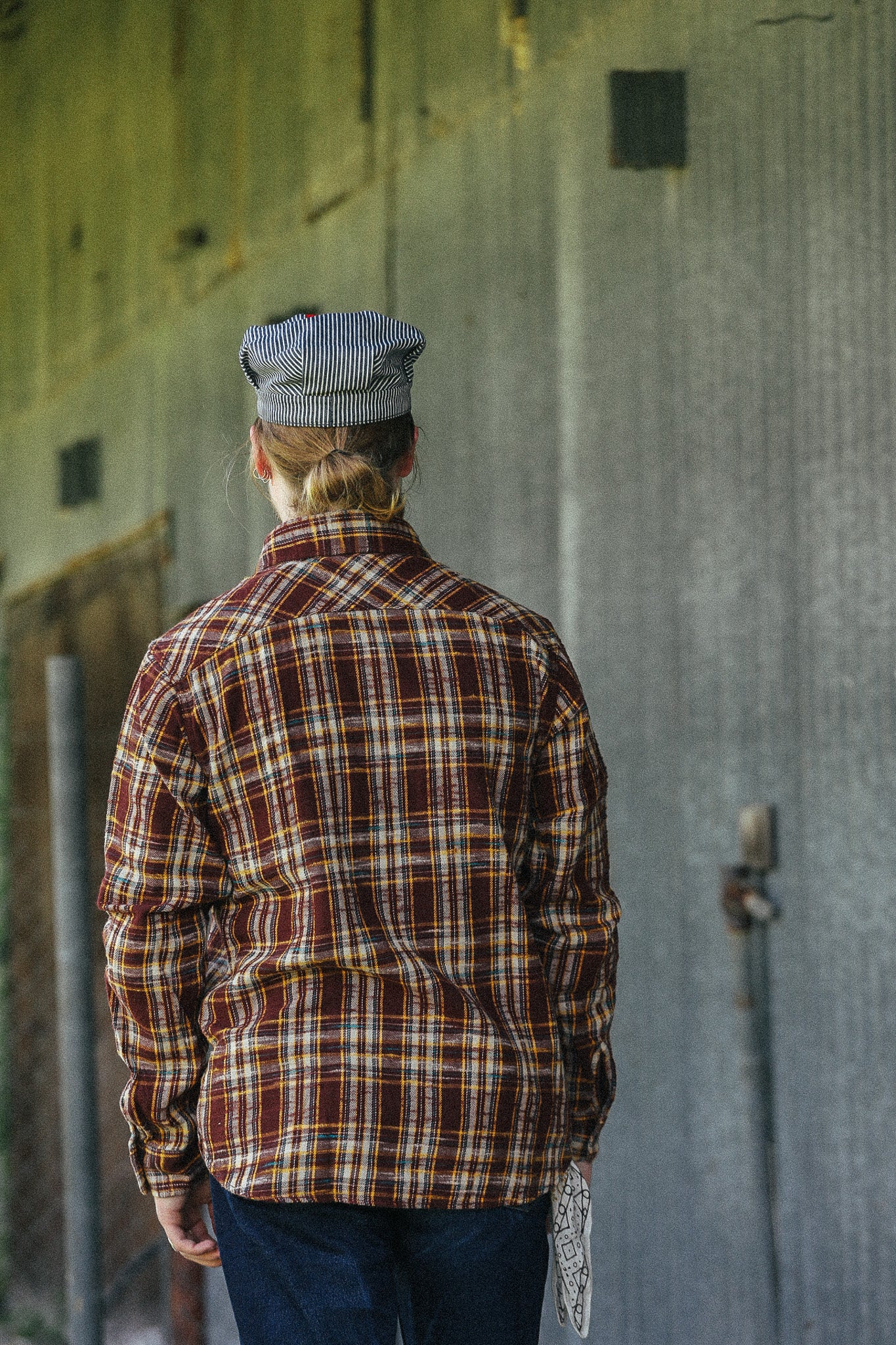 A person viewed from behind wearing a brown and red plaid flannel shirt and a blue-gray striped flat cap, standing against a weathered gray metal wall.
