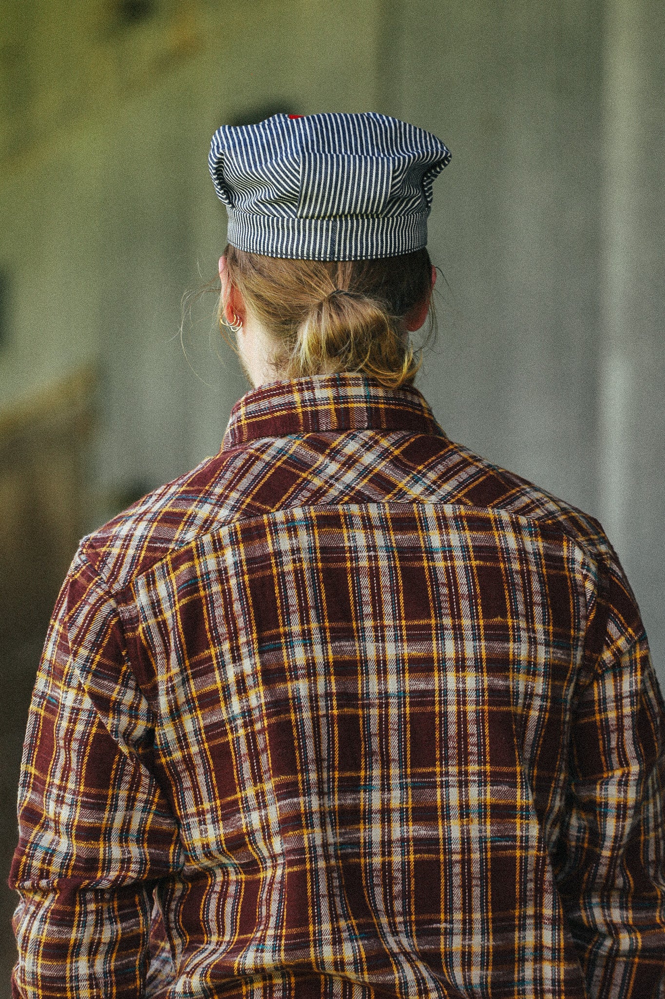 A person viewed from behind wearing a brown and red plaid flannel shirt with yellow and white stripes, and a blue-and-white striped cap, standing against a blurred gray background.