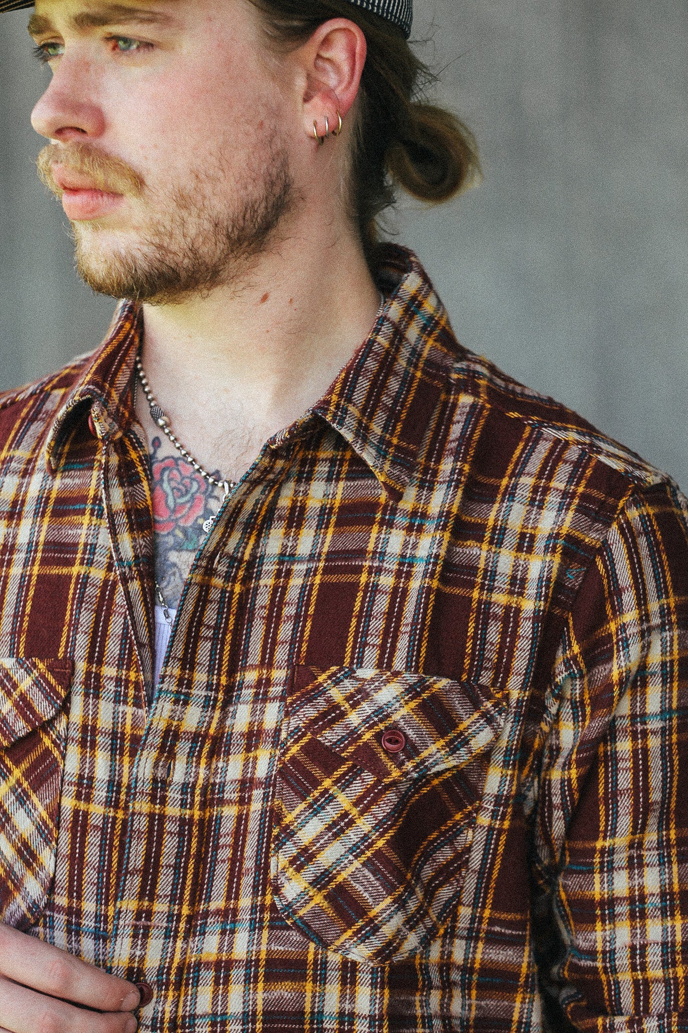 A close-up shot of a man wearing a brown, yellow, and white plaid flannel shirt, looking off to the side against a gray concrete wall.