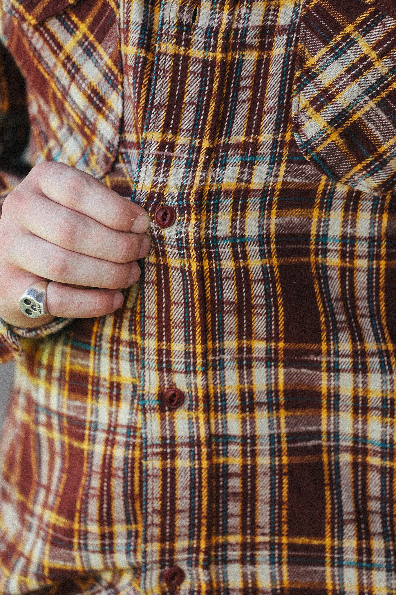 Close-up of a person wearing a brown, yellow, and cream plaid button-up shirt, with a hand pulling the front button.