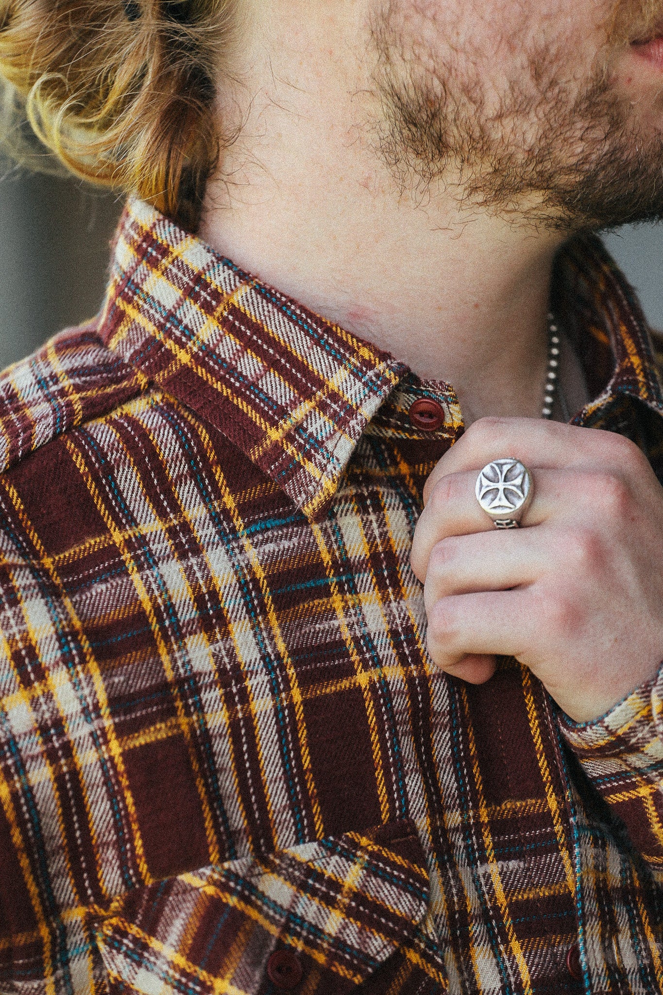 A close-up lifestyle shot of a man wearing a brown, yellow, and white plaid flannel shirt, featuring a detailed silver ring with a cross design on his finger.