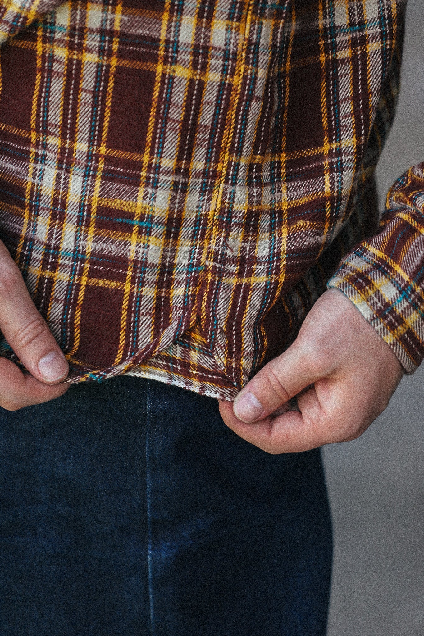 Close-up of a person wearing a brown, yellow, and cream plaid flannel shirt, pulling the hem slightly to show the fabric texture and construction, paired with dark blue jeans.