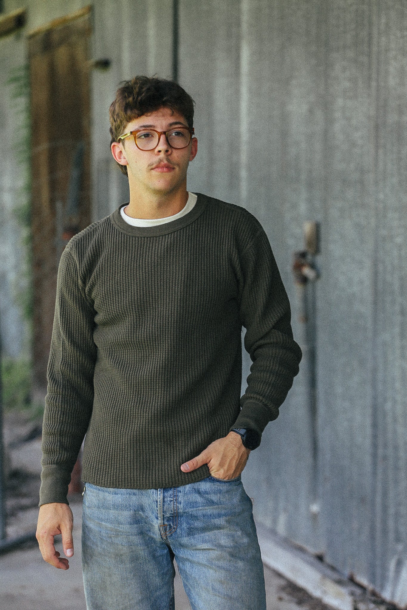 A young man wearing a dark olive green ribbed knit sweater over a white undershirt and blue jeans, standing in front of a weathered gray metal wall.
