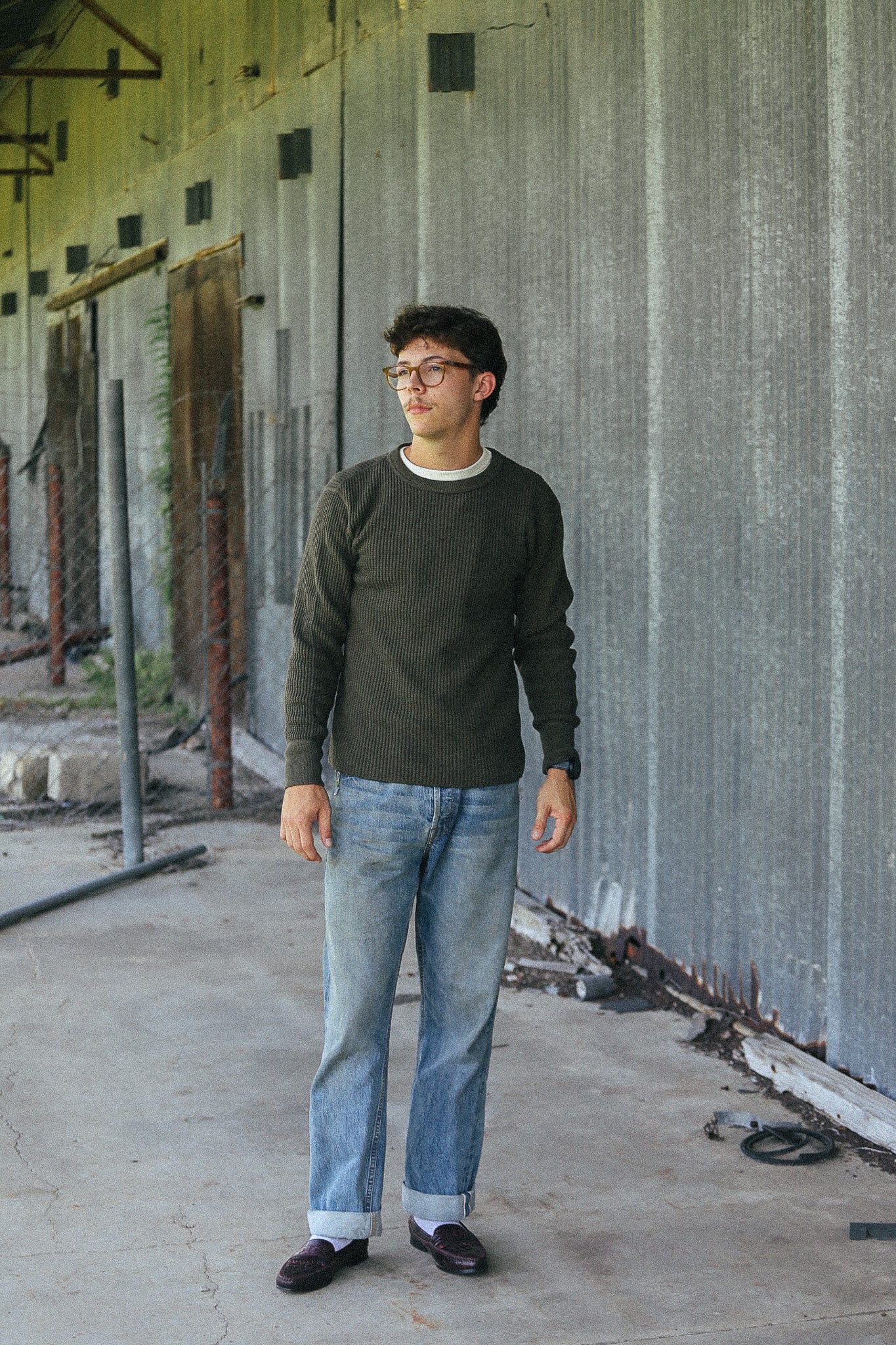 A man wearing a dark olive green long-sleeve crewneck sweater, light blue jeans, white socks, and dark loafers stands in a concrete industrial corridor with a corrugated metal wall.
