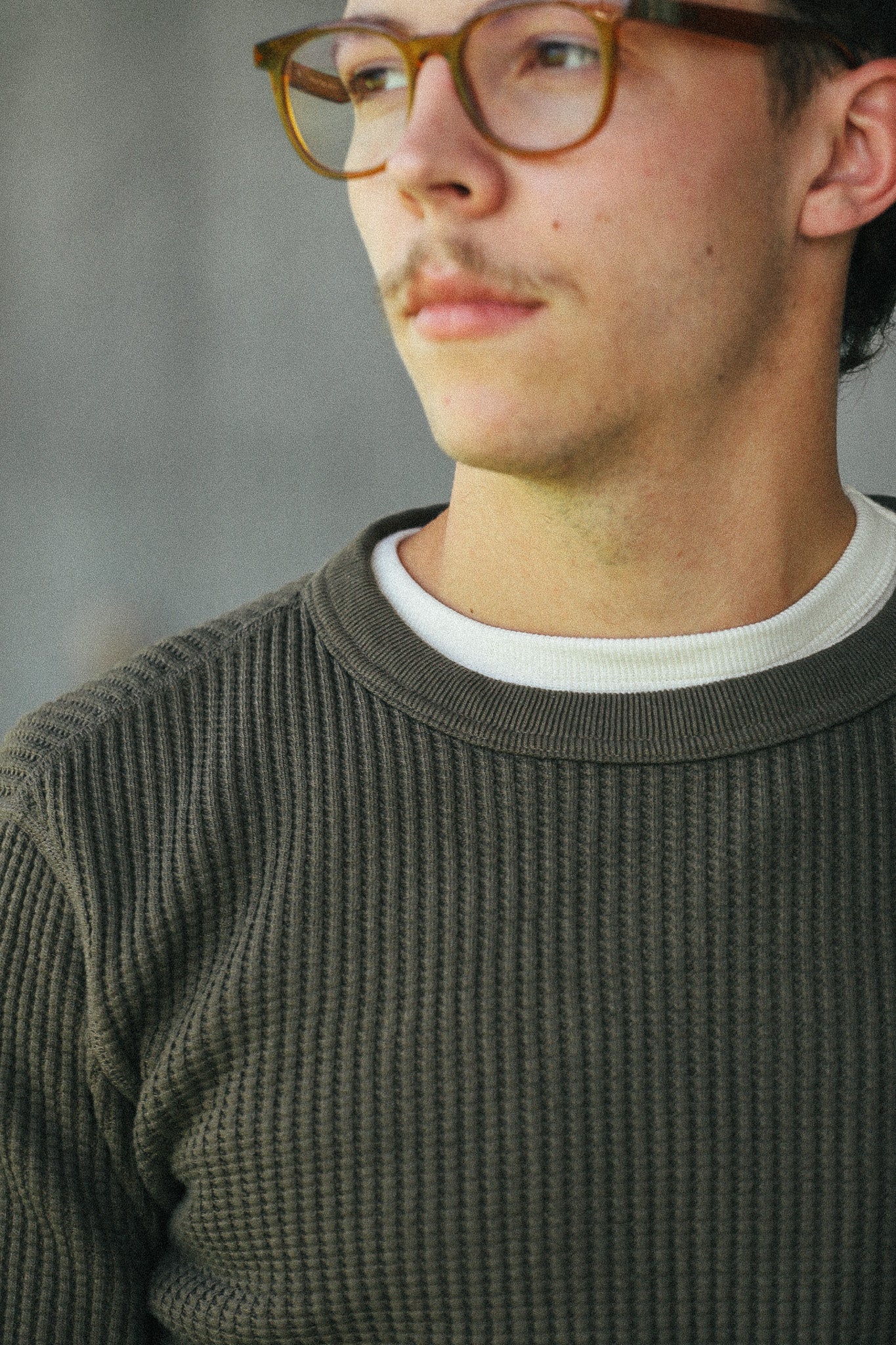 A close-up shot of a man wearing a dark olive green ribbed knit crewneck sweater over a white t-shirt, with brown-rimmed eyeglasses, set against a soft gray background.