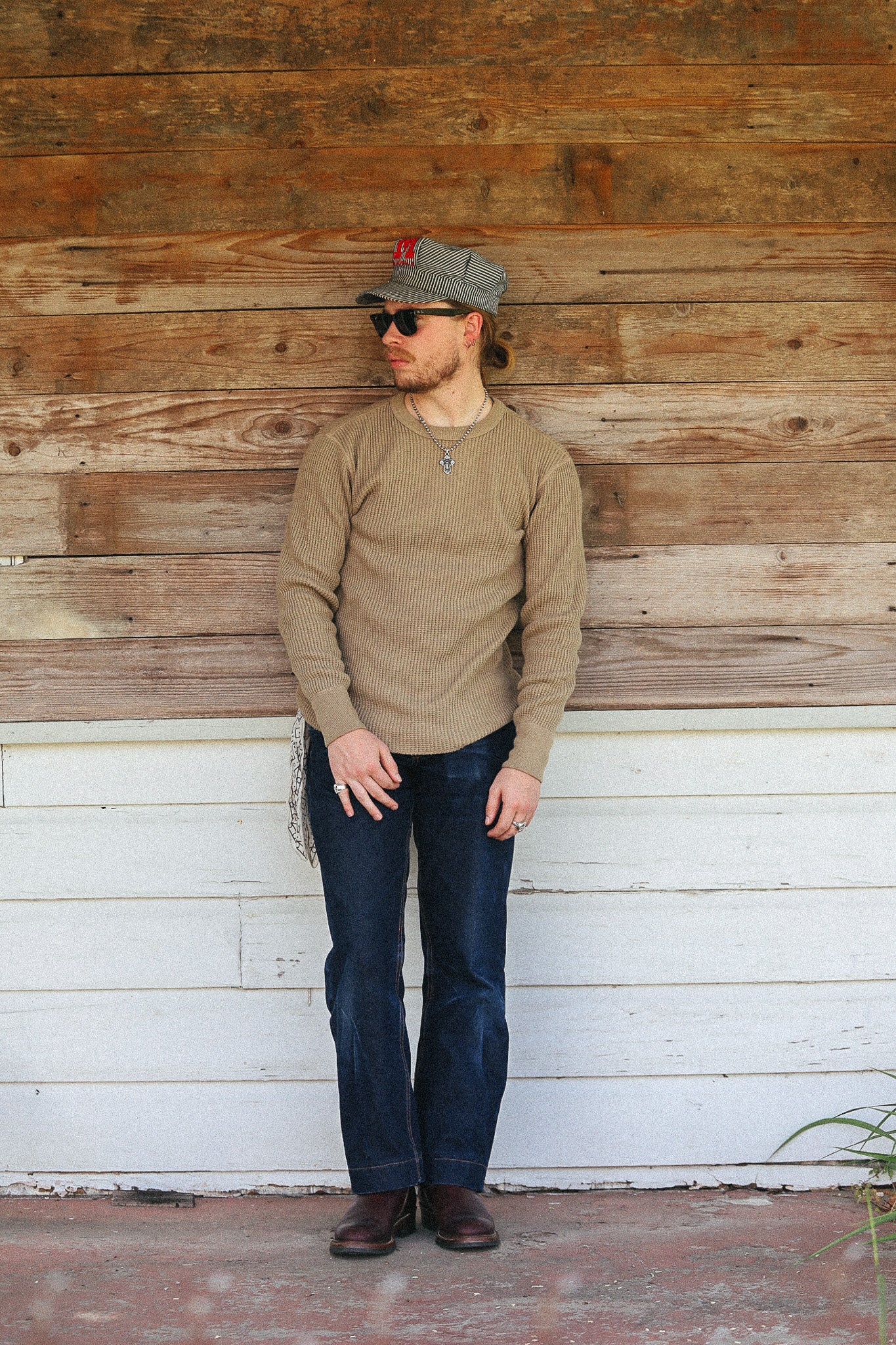 A man stands against a rustic wooden wall wearing a tan long-sleeve knit sweater, dark blue jeans, brown boots, sunglasses, and a gray baseball cap.