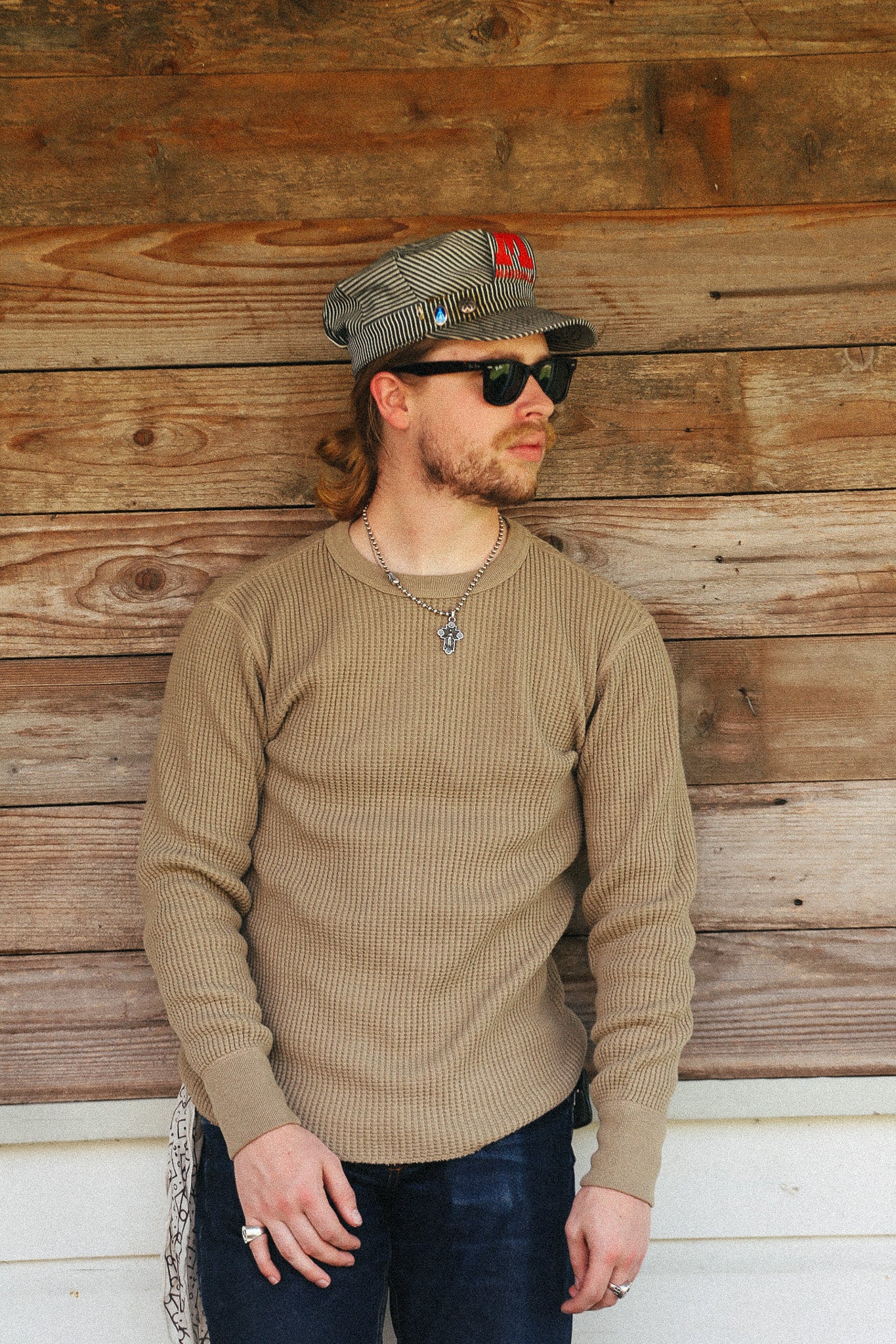 A man wearing a beige ribbed knit crewneck sweater, dark sunglasses, a gray cap, and a silver chain necklace, standing against a rustic wooden wall.
