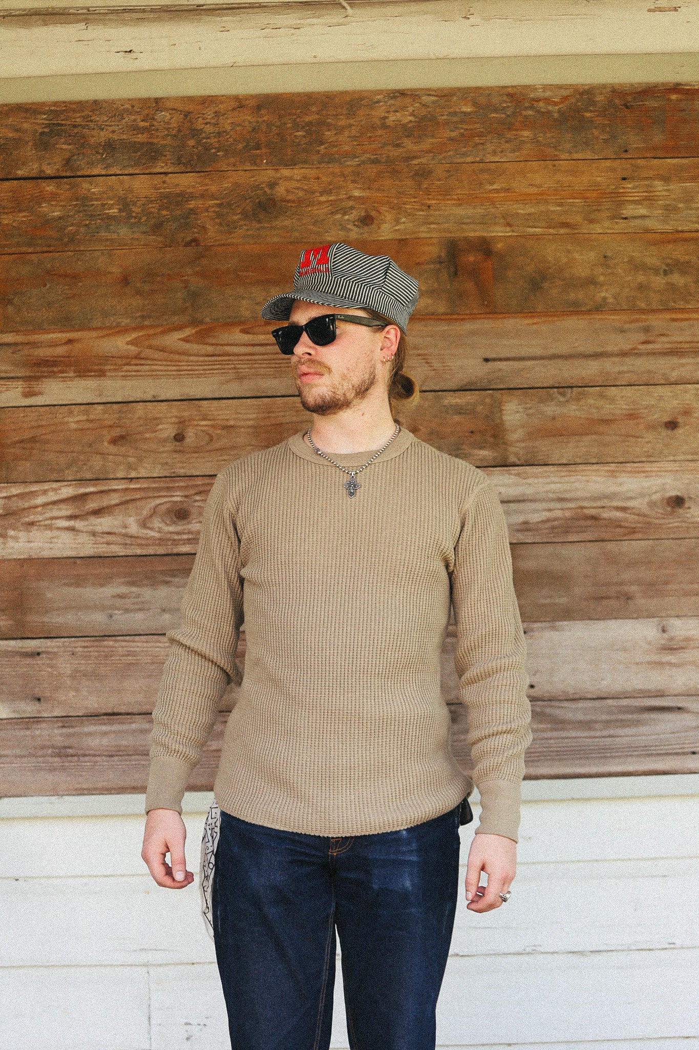A man wearing a beige ribbed long-sleeve sweater, dark blue jeans, a gray baseball cap with a red 'M' logo, and sunglasses, standing in front of a rustic wooden wall.