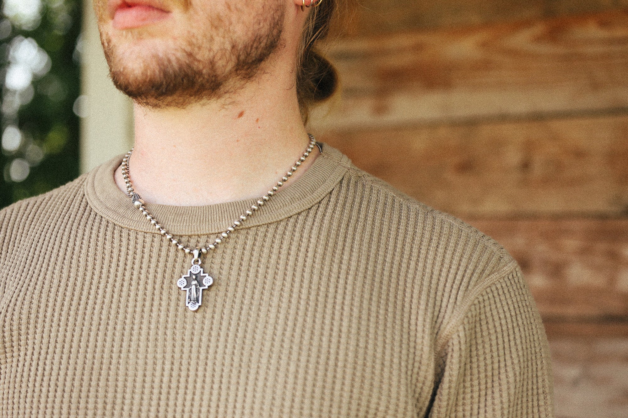 A close-up of a man wearing a silver beaded chain necklace with a silver cross pendant, styled with a beige ribbed knit shirt against a rustic wooden wall background.
