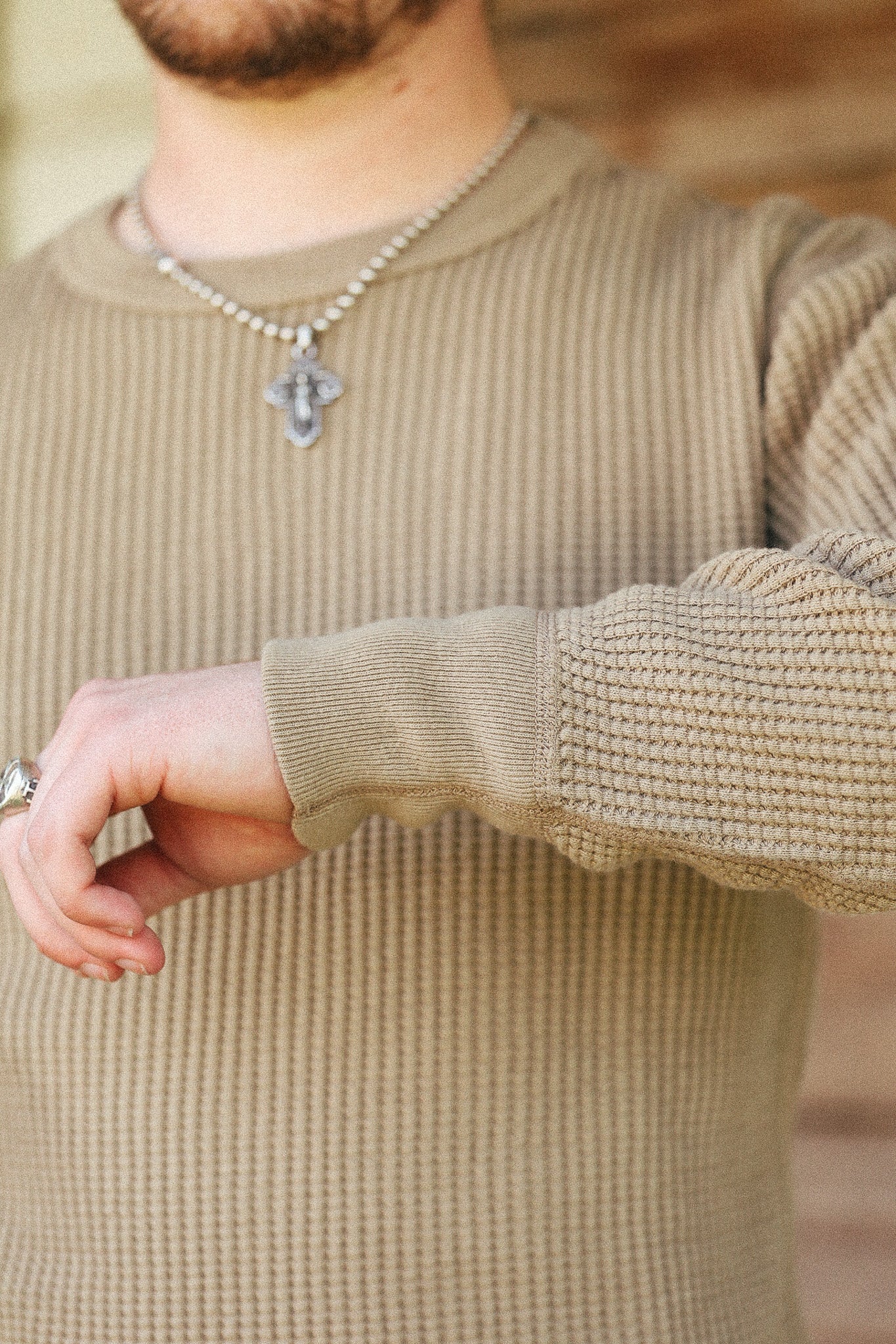 A close-up of a man wearing a beige ribbed knit sweater, a silver beaded necklace with a cross pendant, and a silver ring.