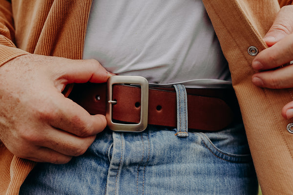 Close-up of a person wearing blue jeans and a brown leather belt with a silver rectangular buckle, adjusting a tan corduroy jacket