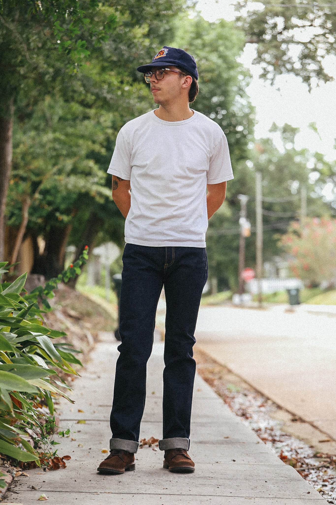 A man standing on a sidewalk outdoors wearing a white crewneck t-shirt, dark blue jeans, brown boots, and a navy baseball cap.