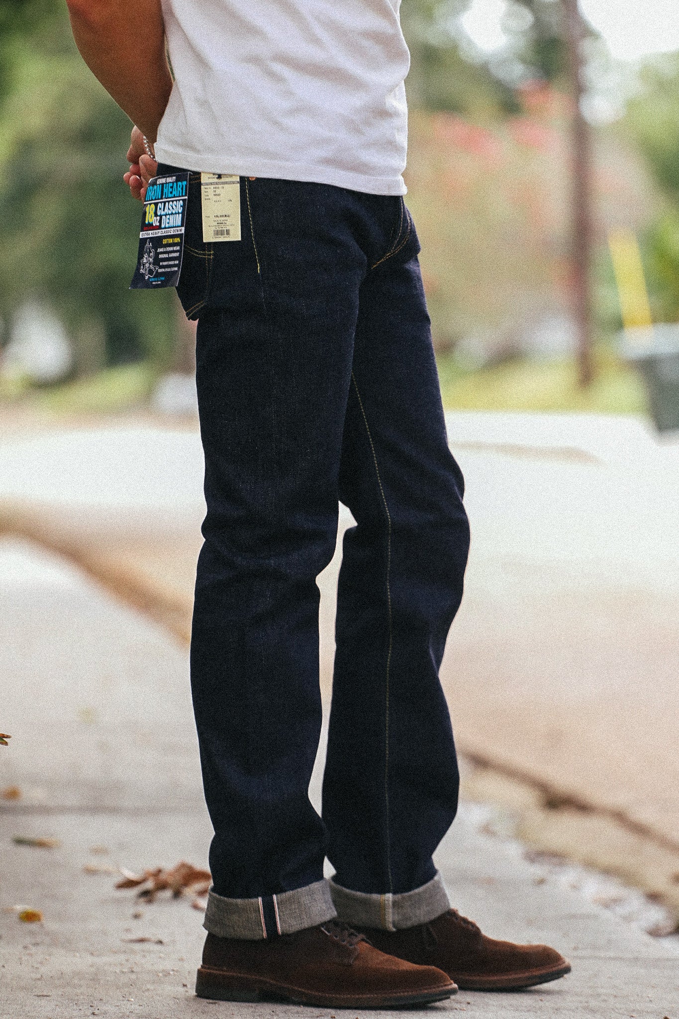 A person outdoors wearing dark indigo denim jeans with rolled cuffs, a white t-shirt, and brown suede boots. A colorful product tag is attached to the back pocket of the jeans.