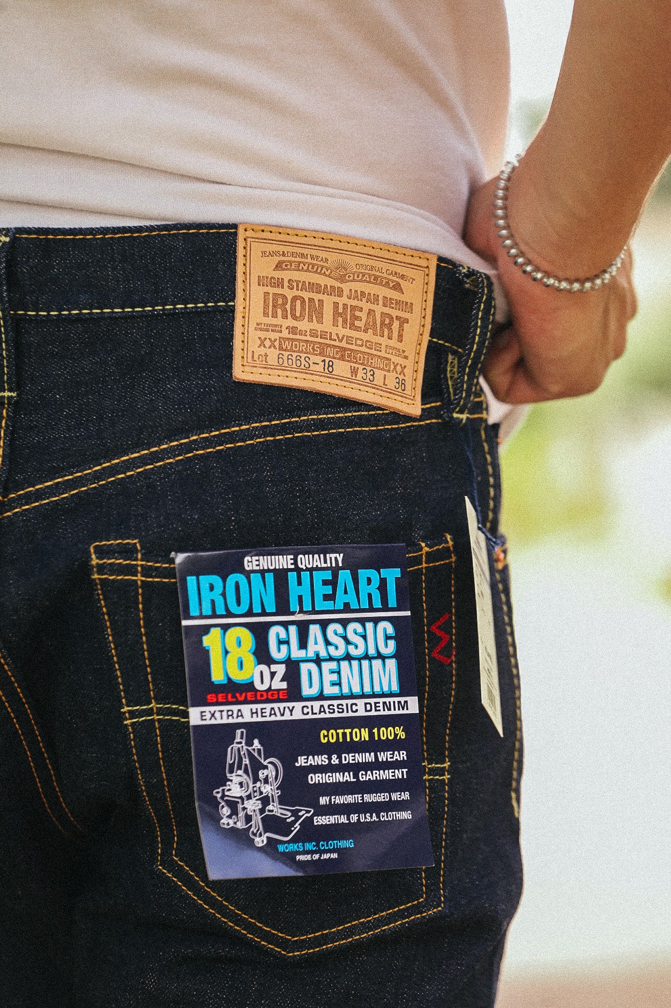 Close-up of the back waistband of dark blue denim jeans worn by a person, showing the tan Iron Heart leather patch and a hanging product tag for 18 oz Classic Denim.