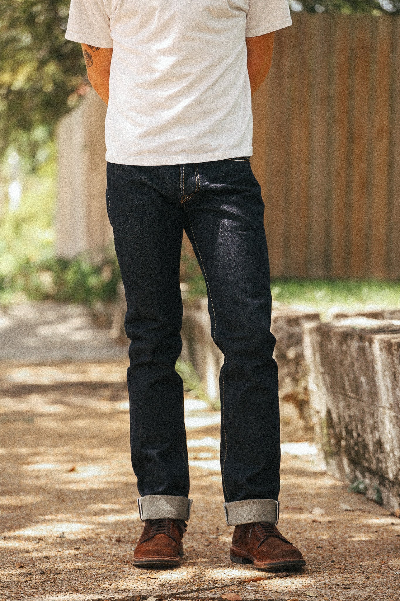 A person standing outdoors wearing dark indigo denim jeans with rolled cuffs, a white t-shirt, and brown leather shoes on a sunlit path.
