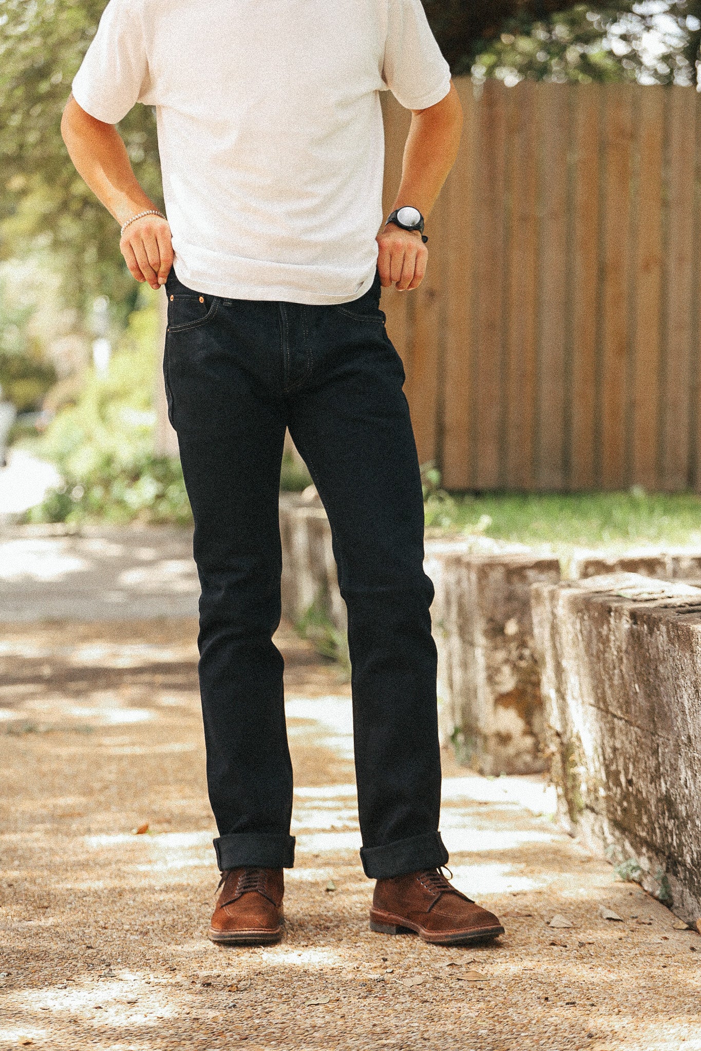 A man wearing dark wash slim-fit jeans, a white t-shirt, and brown leather boots, standing outdoors on a paved path with a wooden fence in the background.
