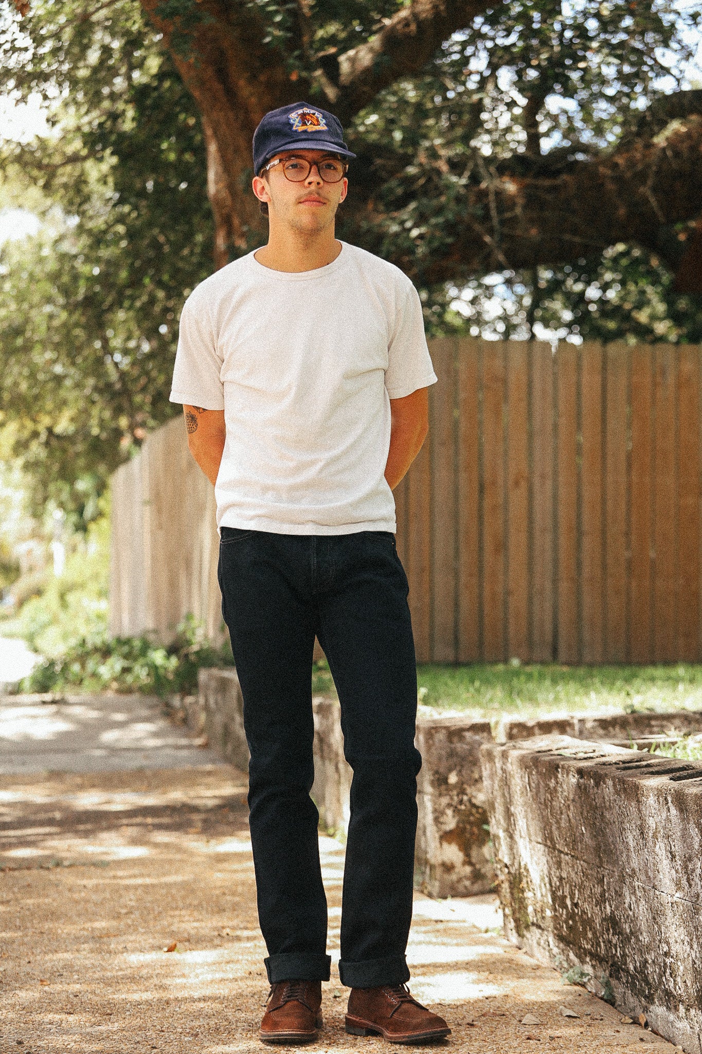 A man standing outdoors on a sunlit path wearing a white crew-neck t-shirt, dark indigo jeans, brown leather shoes, and a navy baseball cap with a red and yellow logo.