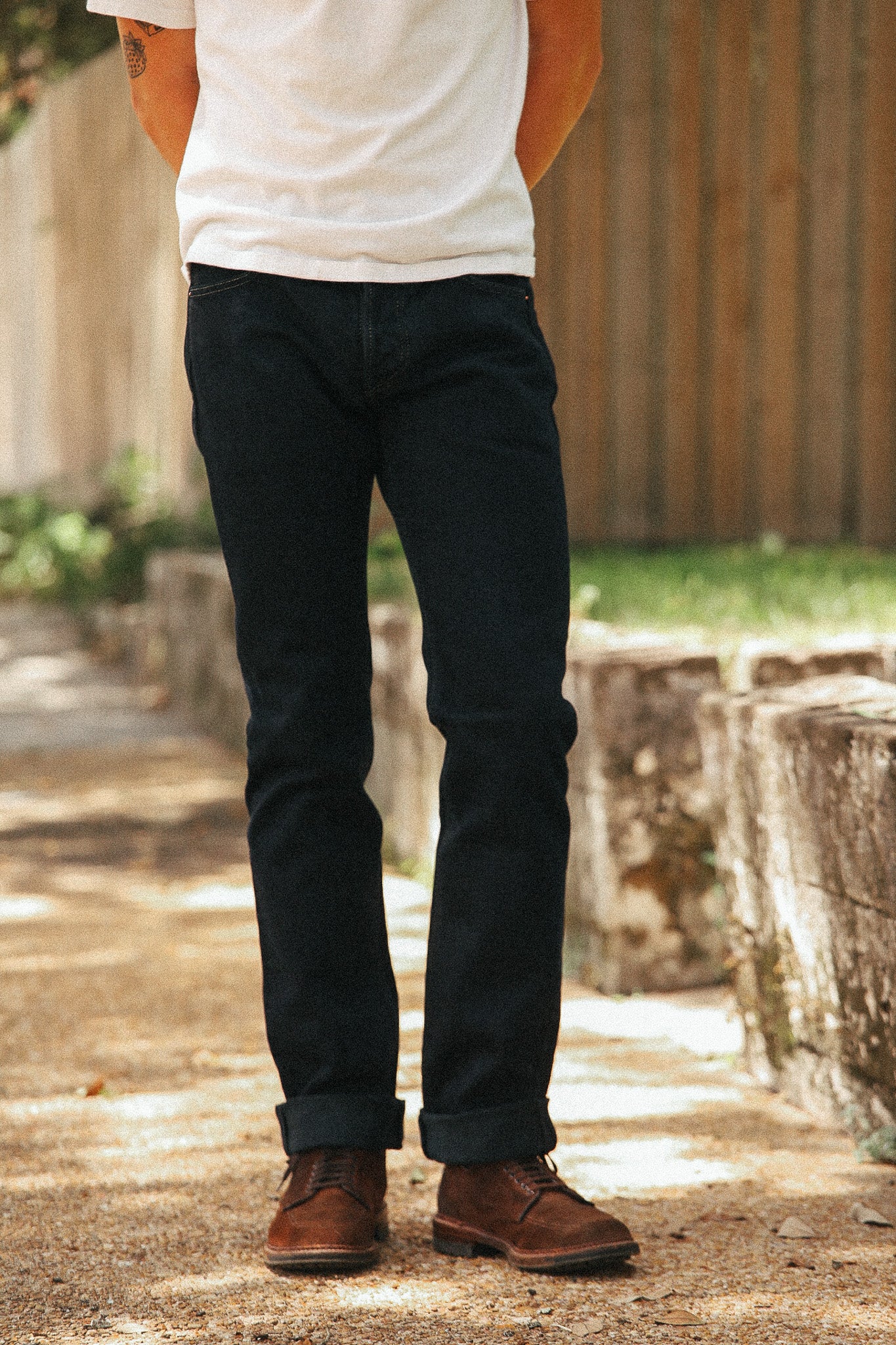 A person standing outdoors wearing dark indigo jeans, a white t-shirt, and brown leather shoes on a sunlit gravel path with a wooden fence in the background.
