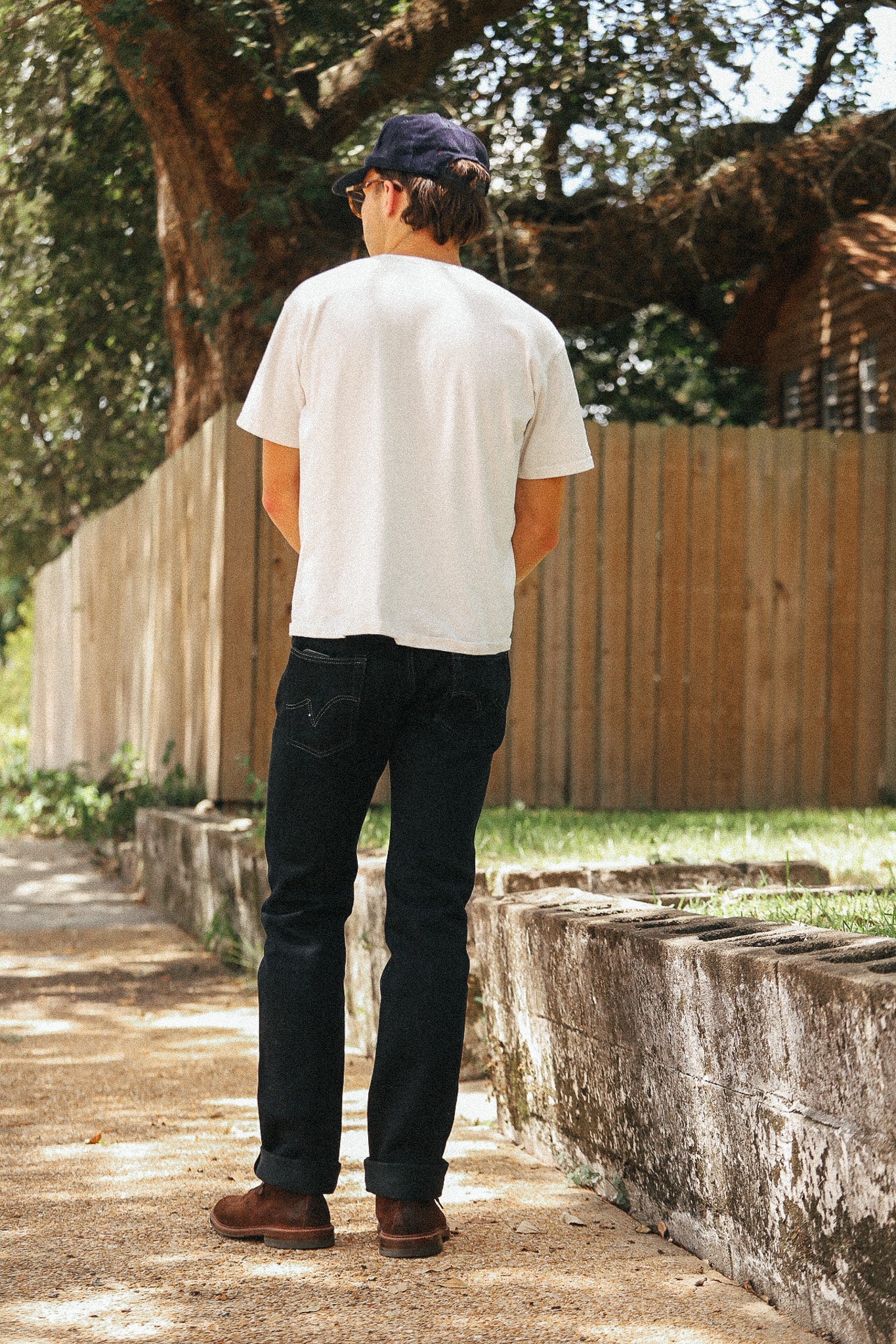 A man standing outdoors on a dirt path, viewed from behind, wearing a white crewneck t-shirt, dark blue jeans, brown boots, and a navy baseball cap. A wooden fence and trees are in the background.