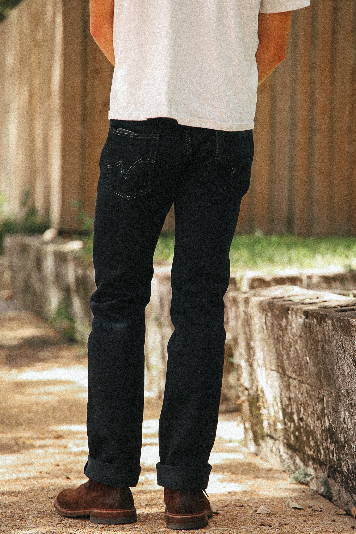 A person shown from the back wearing dark indigo jeans, a white t-shirt, and brown boots, standing outdoors on a stone path with a wooden fence in the background.