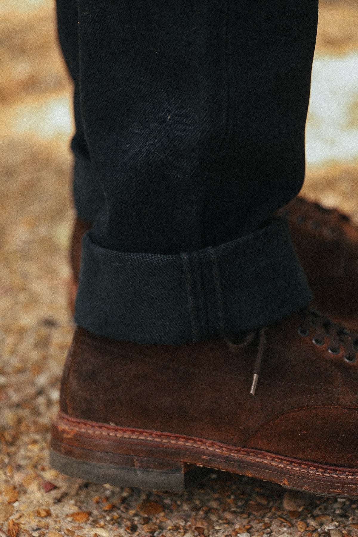 Close-up of a person's lower legs wearing dark charcoal cuffed jeans and brown suede lace-up boots on a gravel surface.