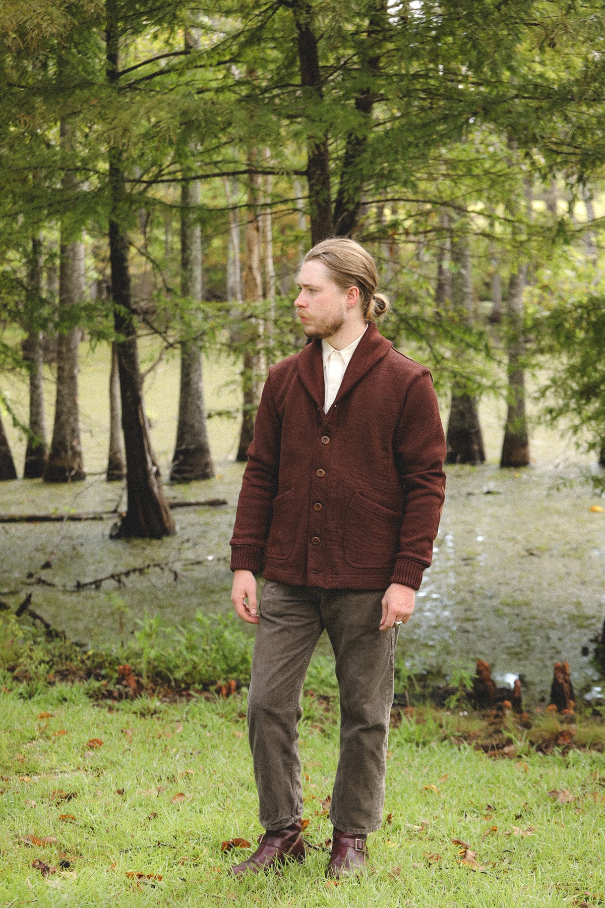 A man standing outdoors in a grassy, tree-filled area wearing a dark brown button-front cardigan over a cream shirt, olive pants, and brown boots.