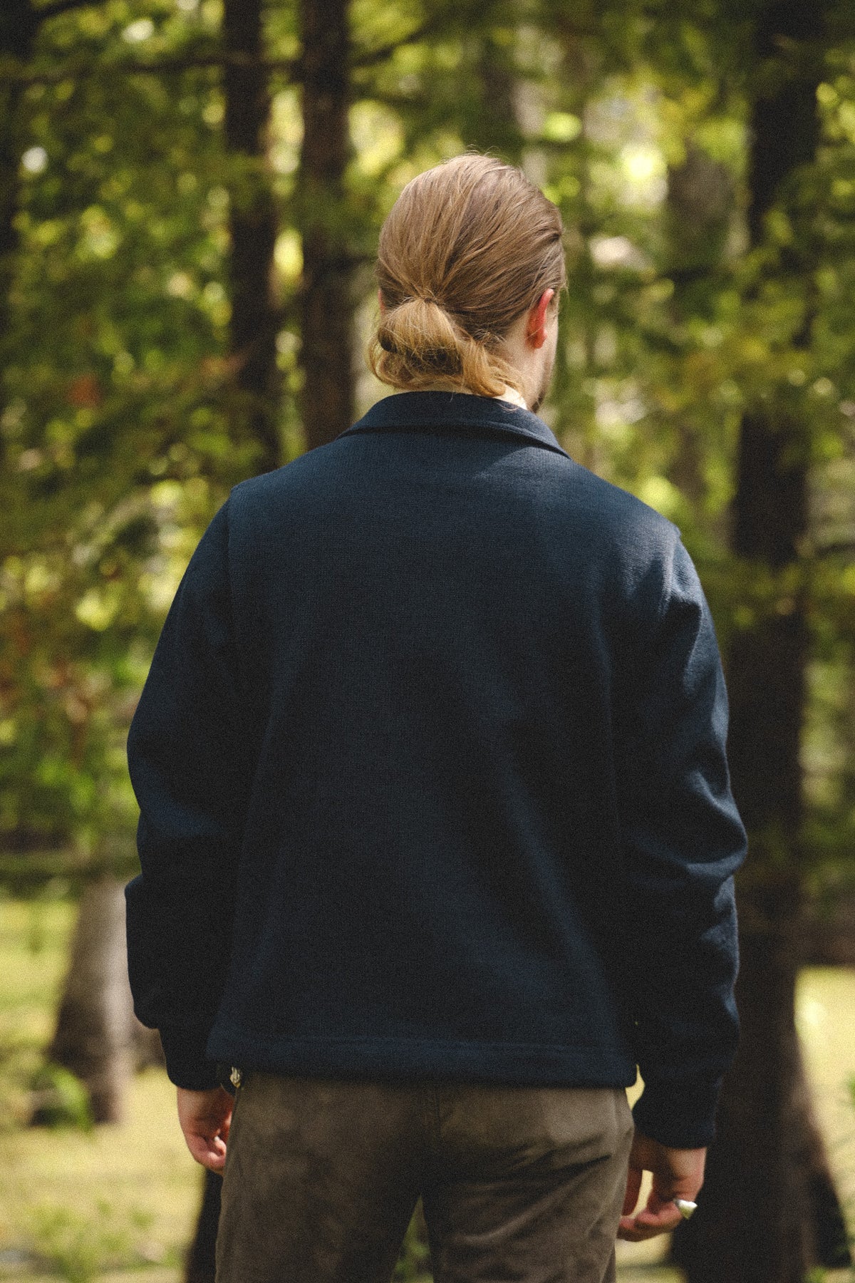 A person viewed from behind wearing a dark navy fleece jacket and brown pants, standing in a sunlit forest with trees in the background.