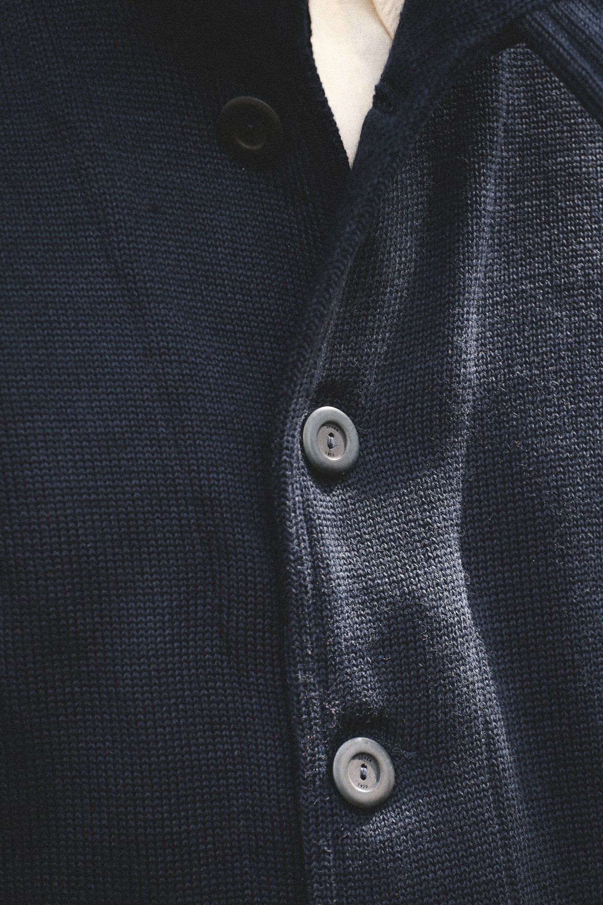 Close-up of a dark navy textured knit button-front garment, showing the fabric weave and gray buttons, with a small sliver of white inner collar visible.