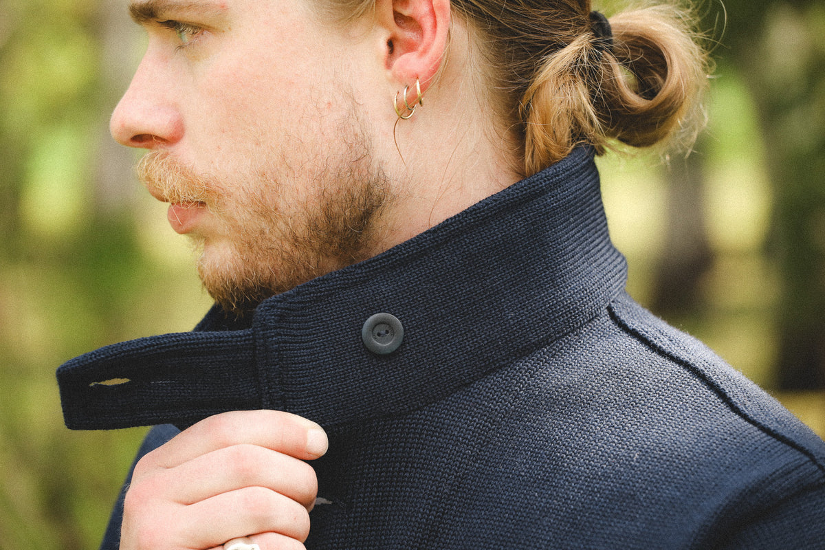 Close-up side profile of a man outdoors wearing a dark navy textured coat with a high ribbed collar and a button closure.