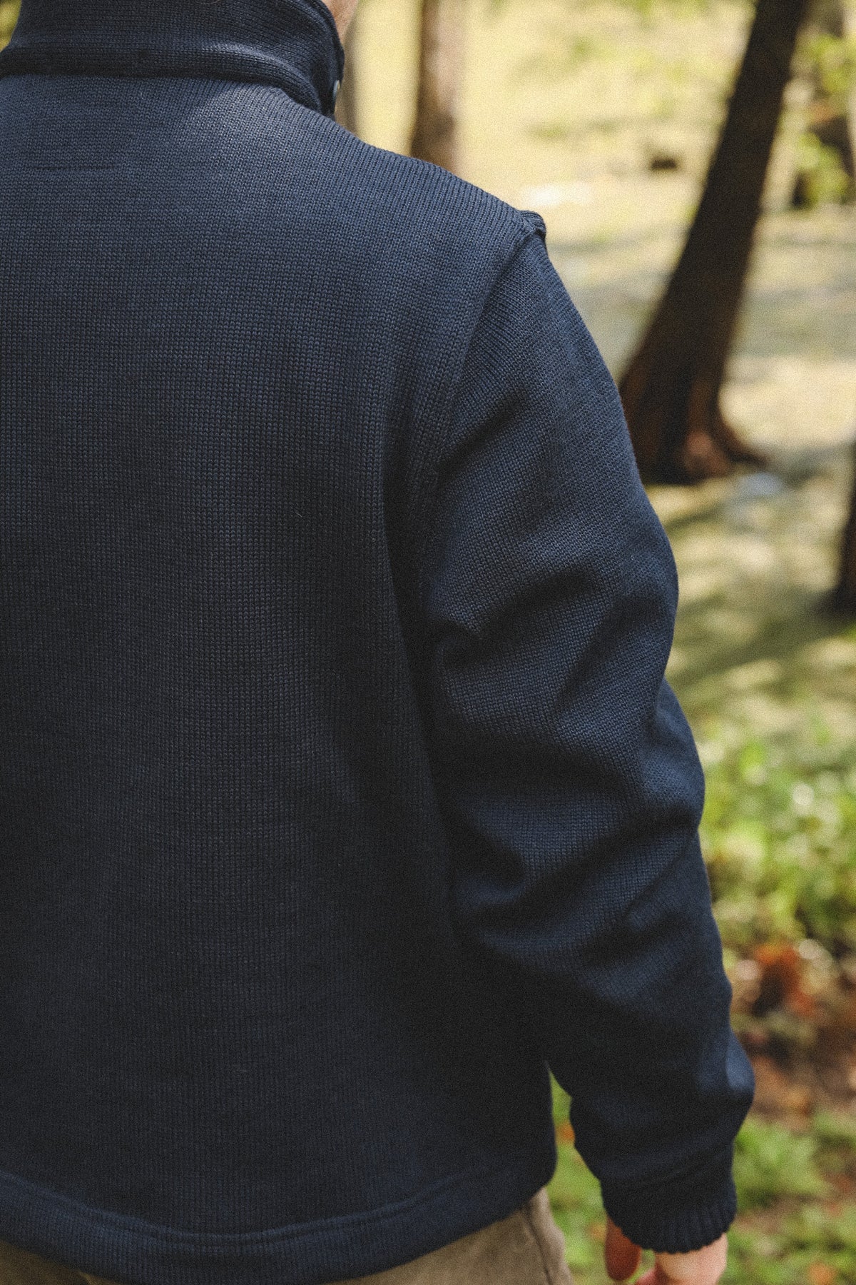 A person outdoors wearing a dark navy ribbed knit quarter-zip fleece jacket, shown from the back with a blurred forest background.