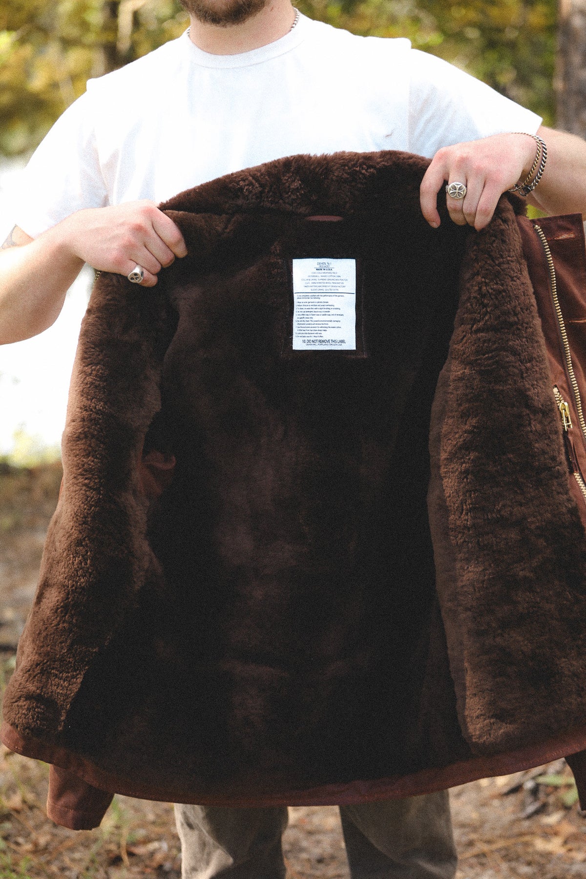 A person outdoors holding open a dark brown shearling-lined jacket to show the interior lining and a white interior label.