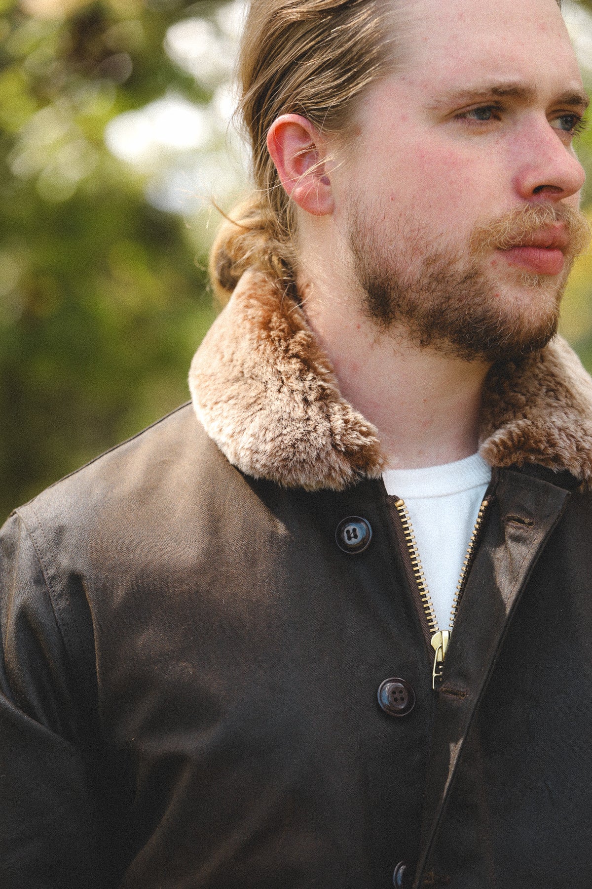 A close-up shot of a man wearing a dark brown waxed canvas jacket with a thick, warm brown shearling collar, layered over a white t-shirt. The background is a soft, out-of-focus natural green bokeh.
