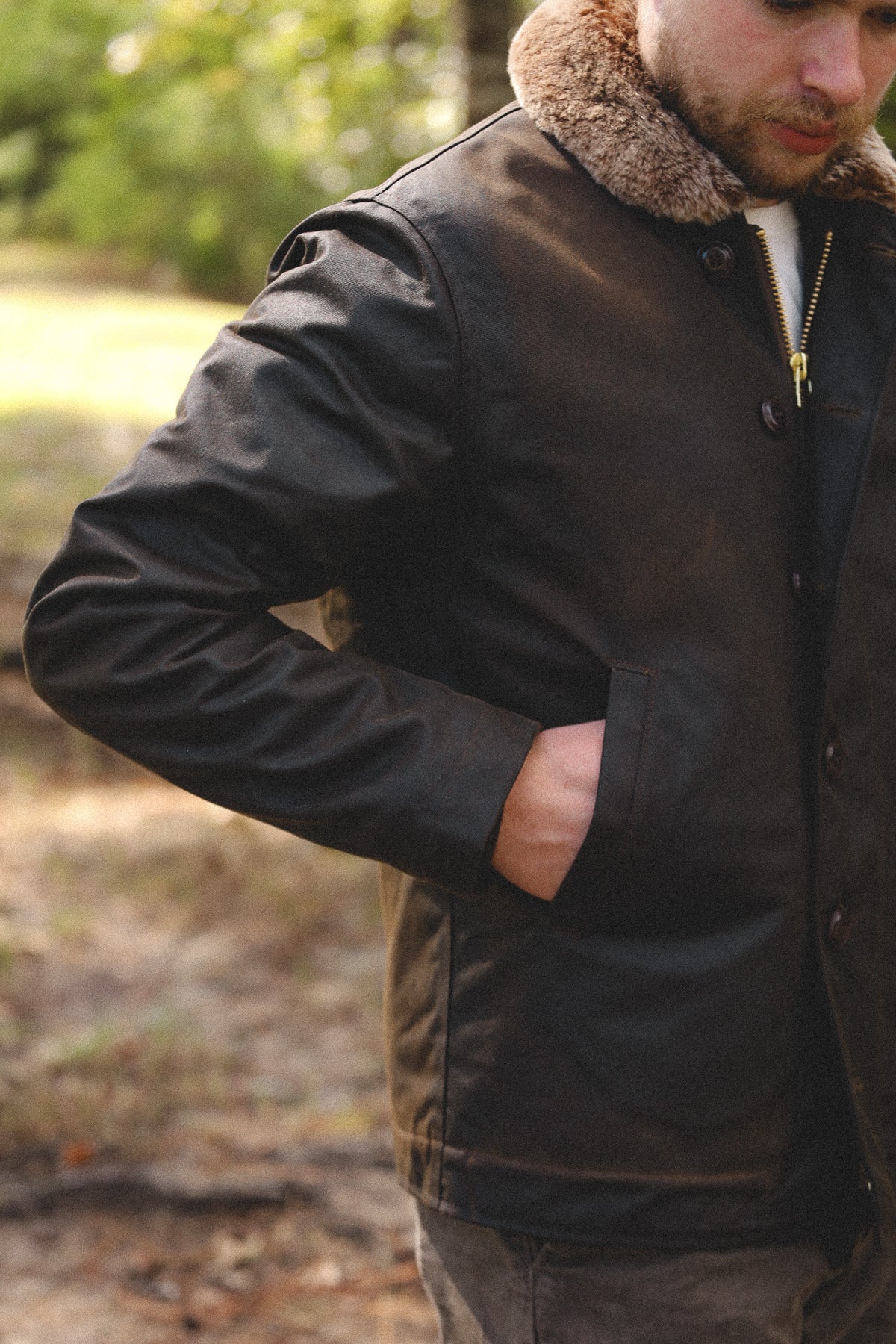 A man wearing a dark brown waxed cotton jacket with a brown shearling collar, standing outdoors in a wooded setting.