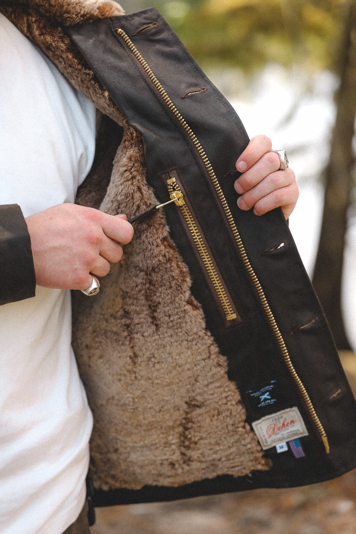 Close-up of a person wearing a dark brown jacket with a thick brown shearling lining, holding the jacket open to show the interior and a gold zipper pocket.