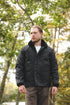 A man wearing a black corduroy jacket with a dark collar, standing outdoors in front of a blurred background of trees.