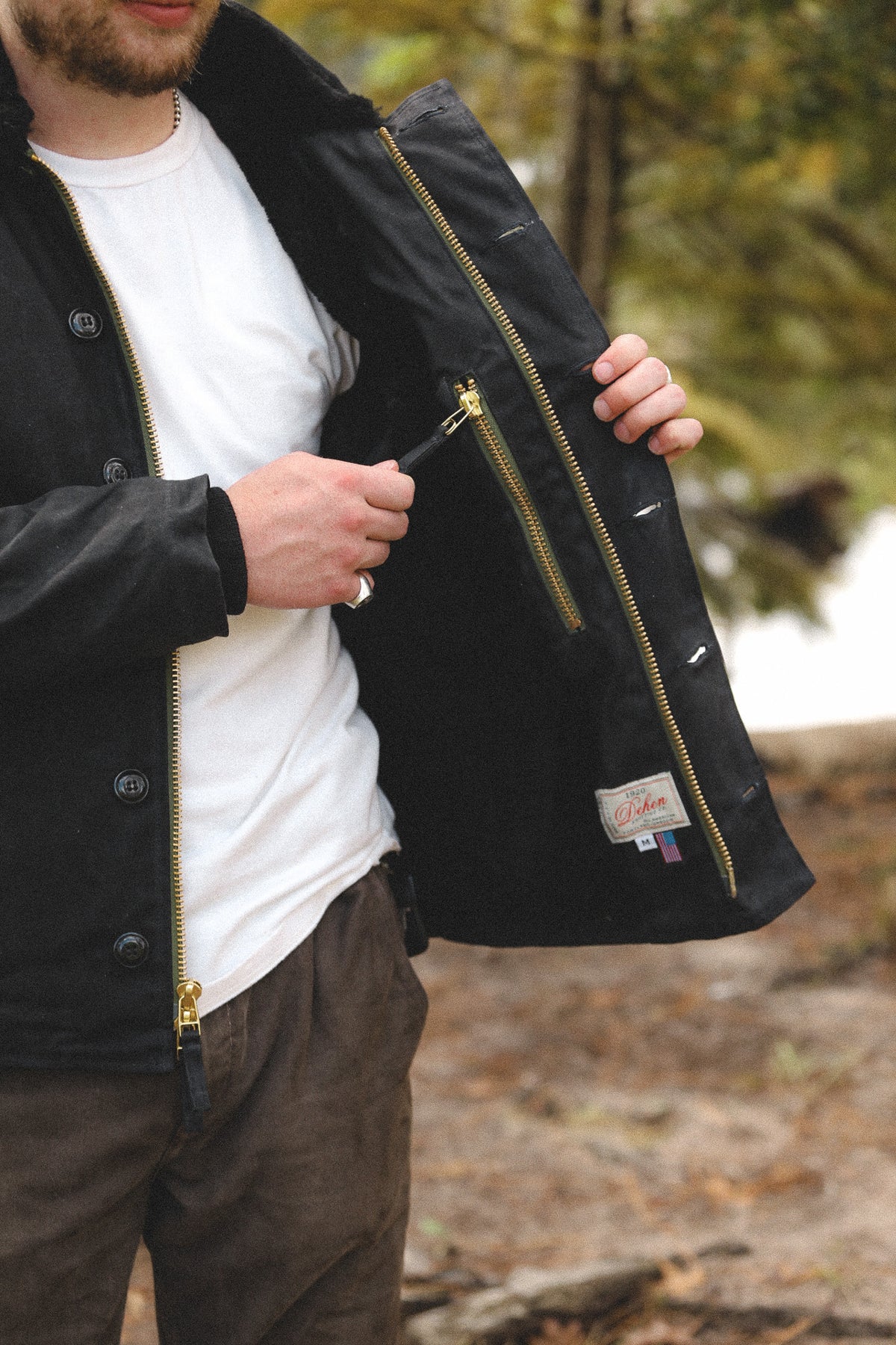 A man outdoors wearing a black waxed jacket over a white t-shirt and brown pants, holding the jacket open to show the interior lining and gold-tone zippers.