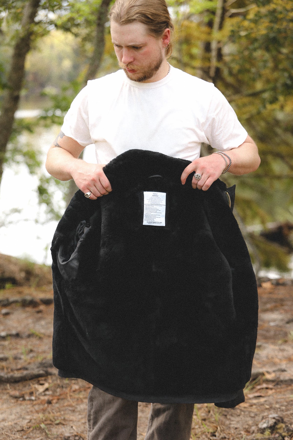 A man in a white t-shirt and gray pants stands outdoors near a wooded area by a body of water, holding up a large black fleece-lined car seat cover with a white product tag attached.