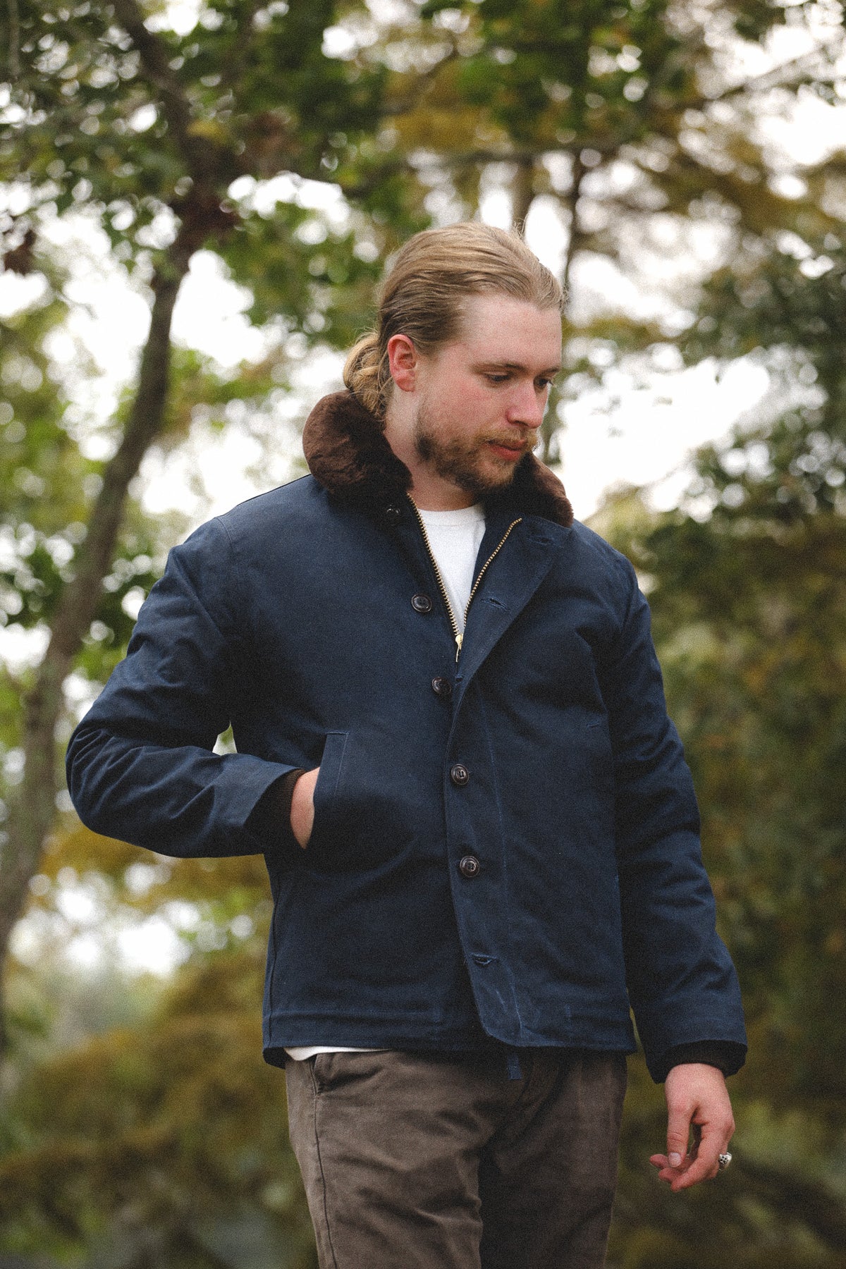 A man wearing a navy blue corduroy jacket with a brown faux-fur collar, standing outdoors in front of a blurred background of trees.