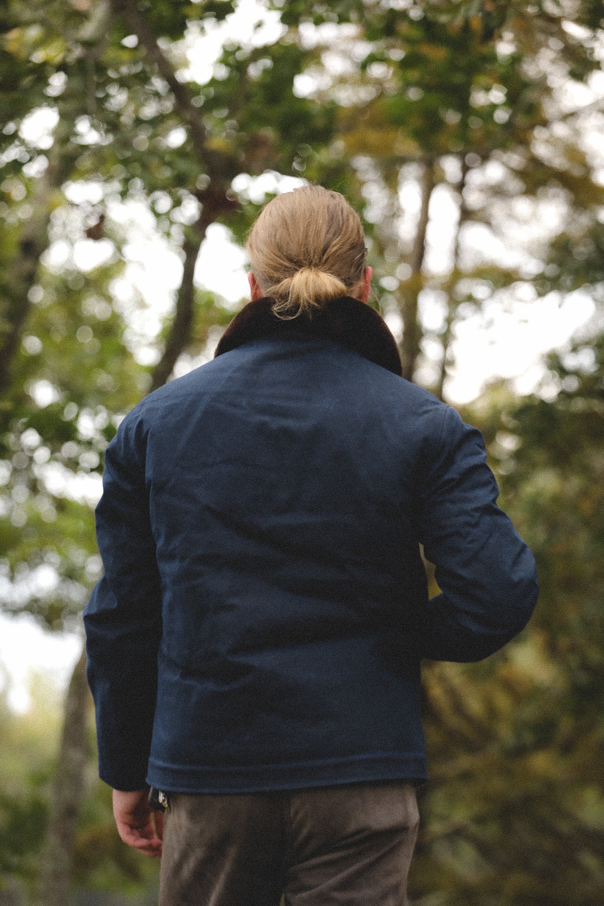 A man viewed from behind wearing a navy blue quilted jacket with a dark collar, standing outdoors in a wooded setting.